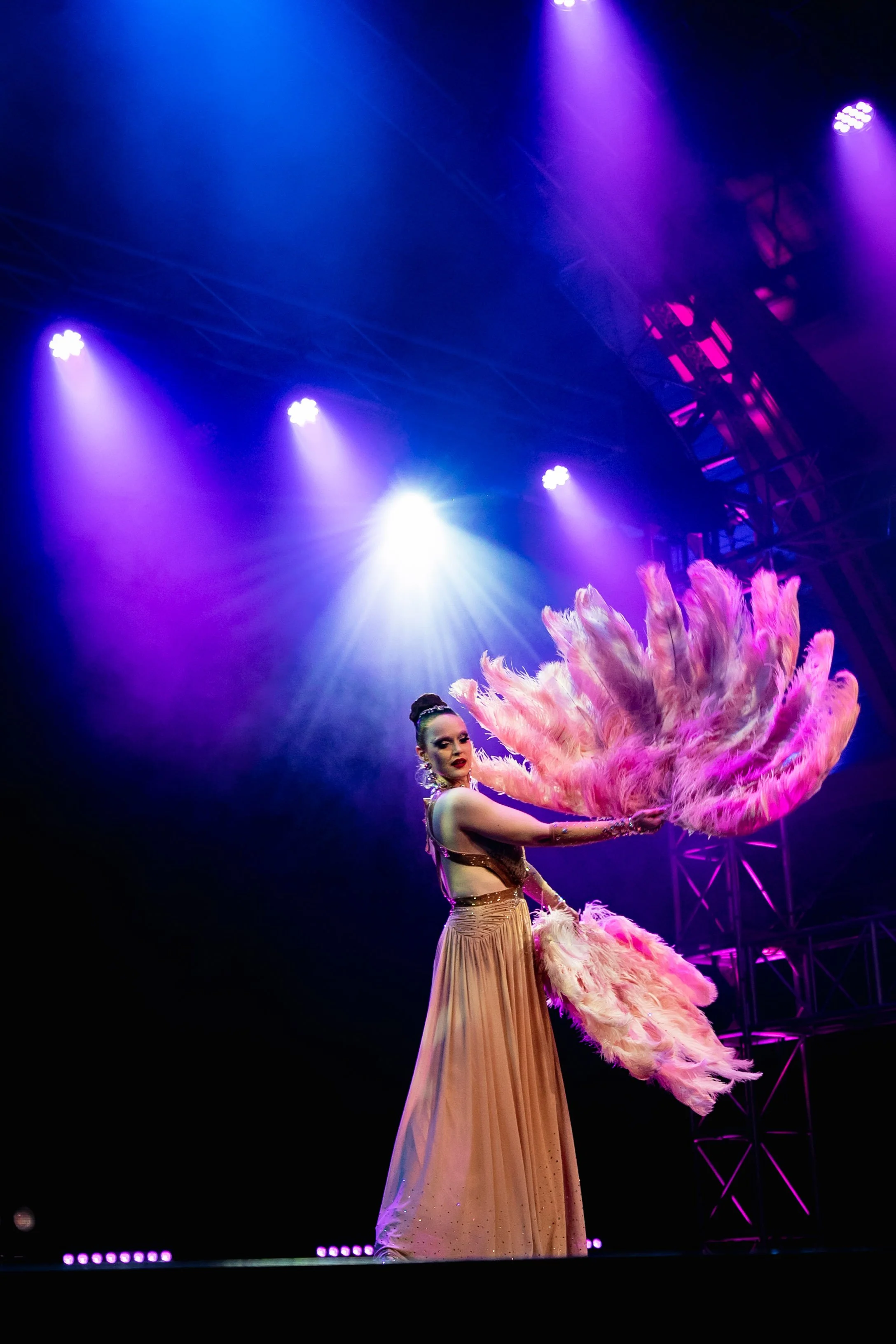 Performer on stage wearing a long, beige skirt and holding a large pink feather fan with stage lighting and purple and blue lights above.