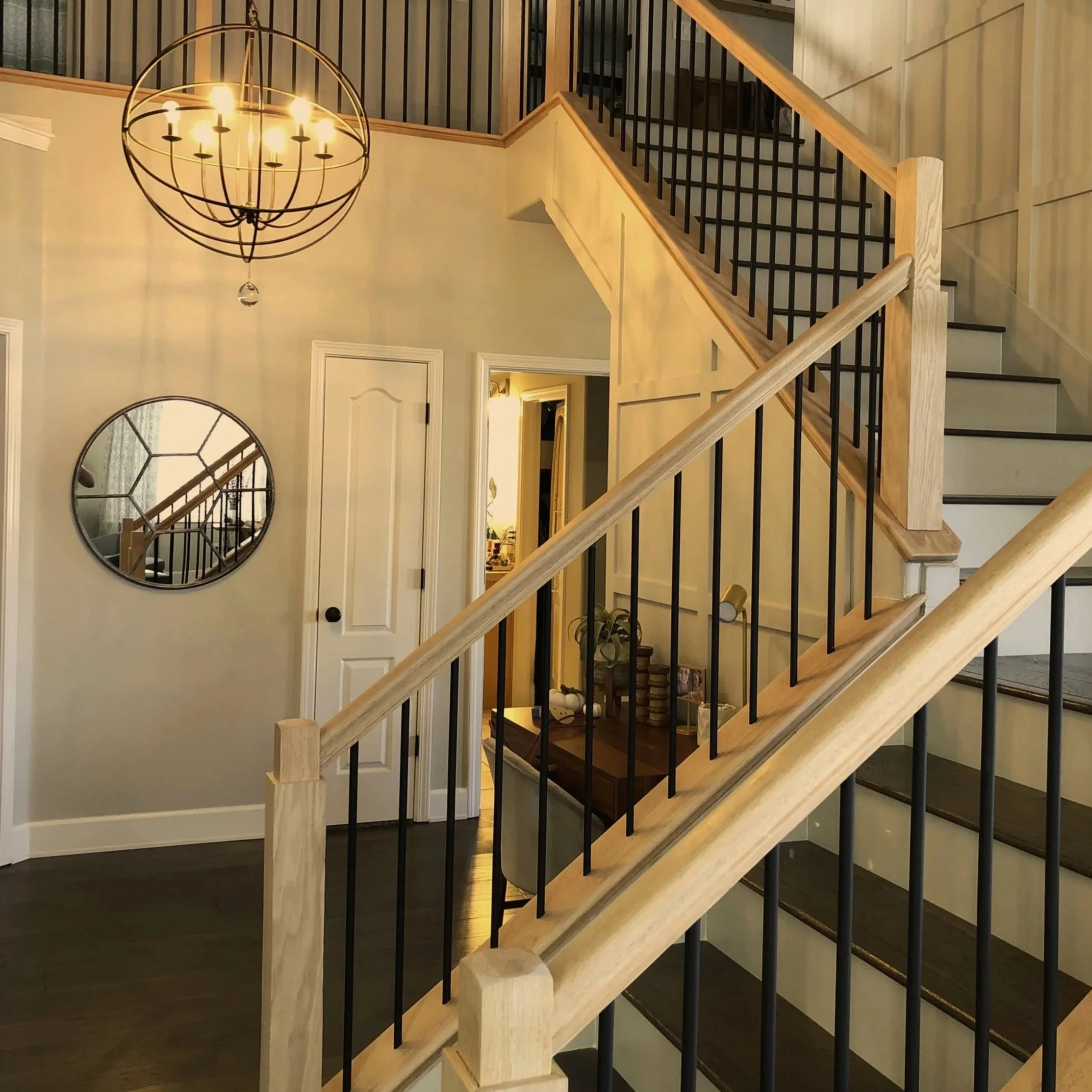 Interior view of a house featuring a wooden staircase with black railings, a chandelier hanging from the ceiling, a round mirror on the wall, and an open door leading to another room.