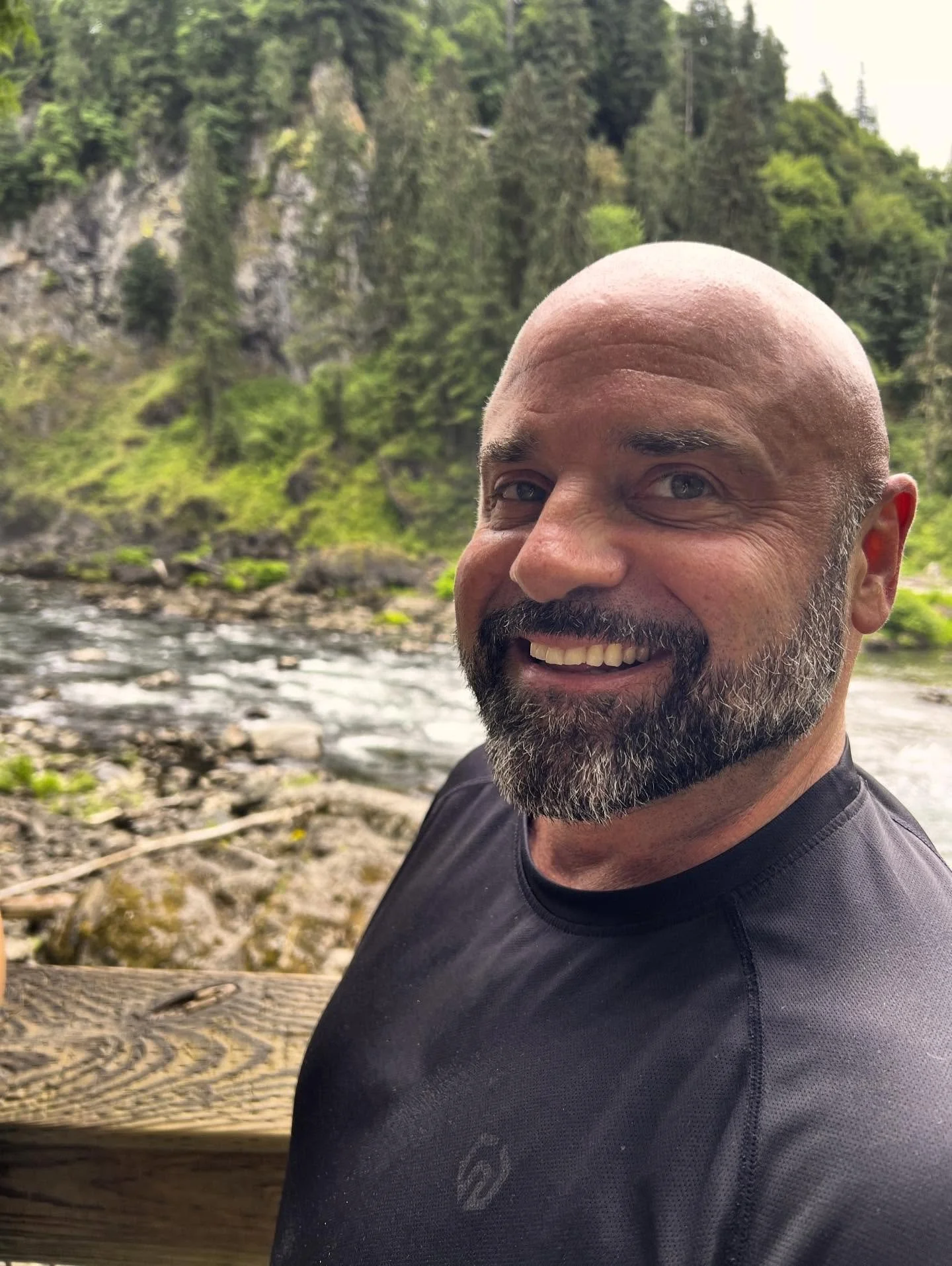 A smiling man with a beard and bald head outdoors near a river, with a lush green forest and rocky cliffs in the background.
