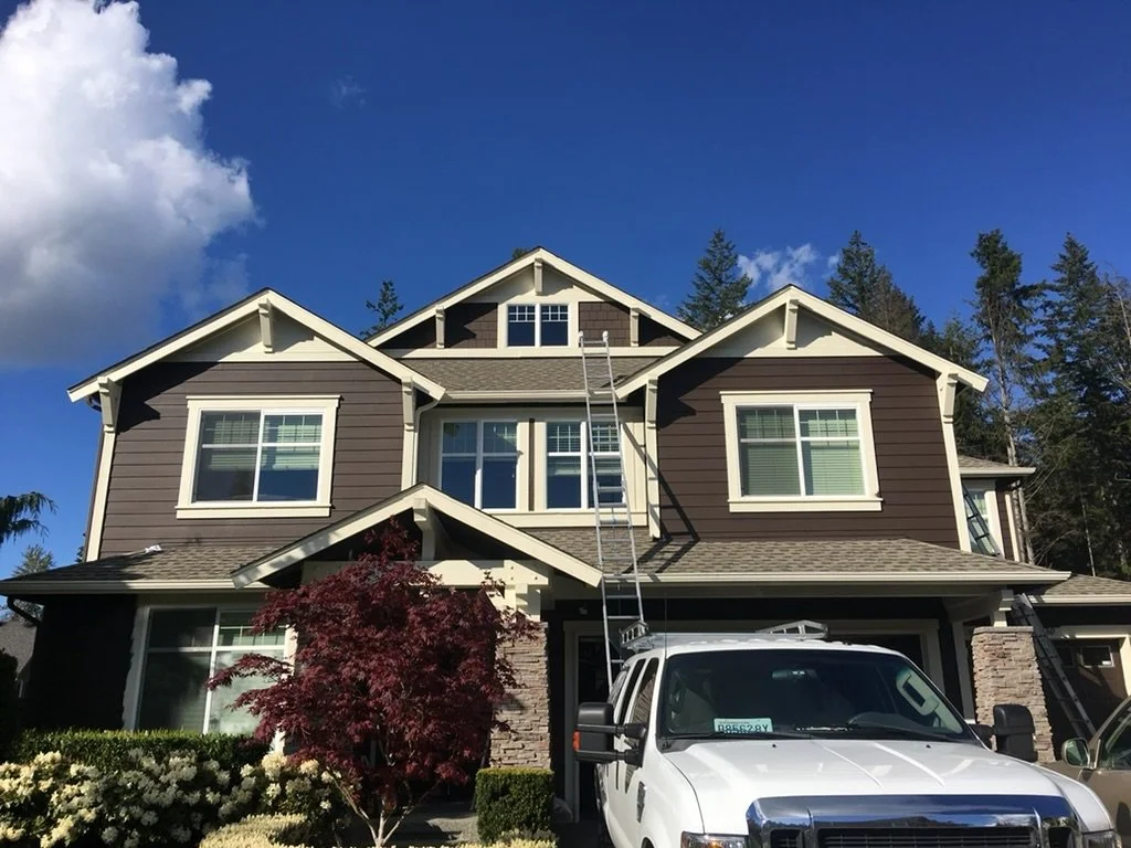 A large two-story house with brown horizontal siding, white trim, and multiple windows, with a ladder leaning against it and a white van parked in the driveway. There are trees and a blue sky with clouds in the background.