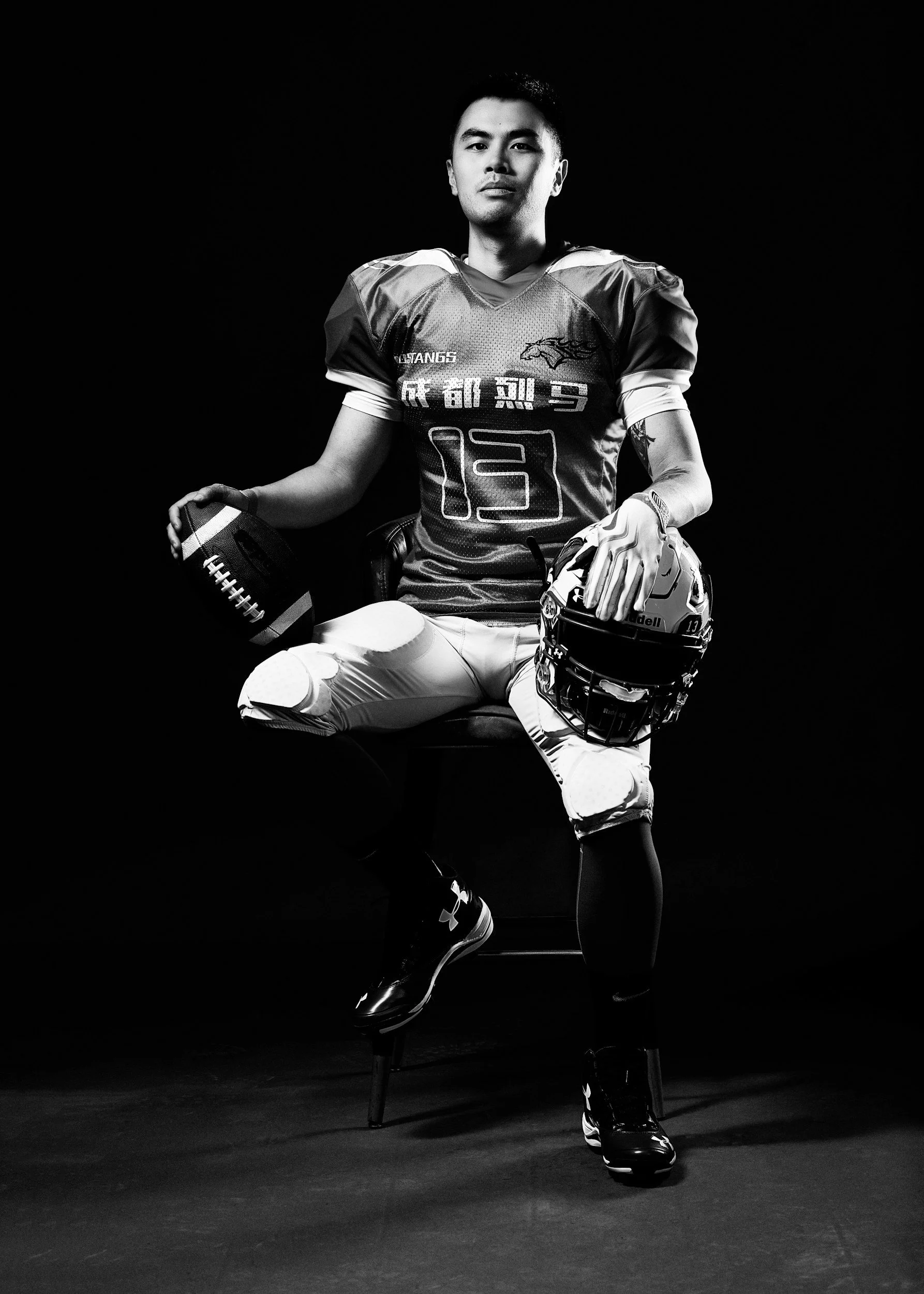 Black and white photo of a football player sitting on a chair, holding a football in his right hand and a helmet in his left hand, wearing a football uniform and cleats, against a dark background.
