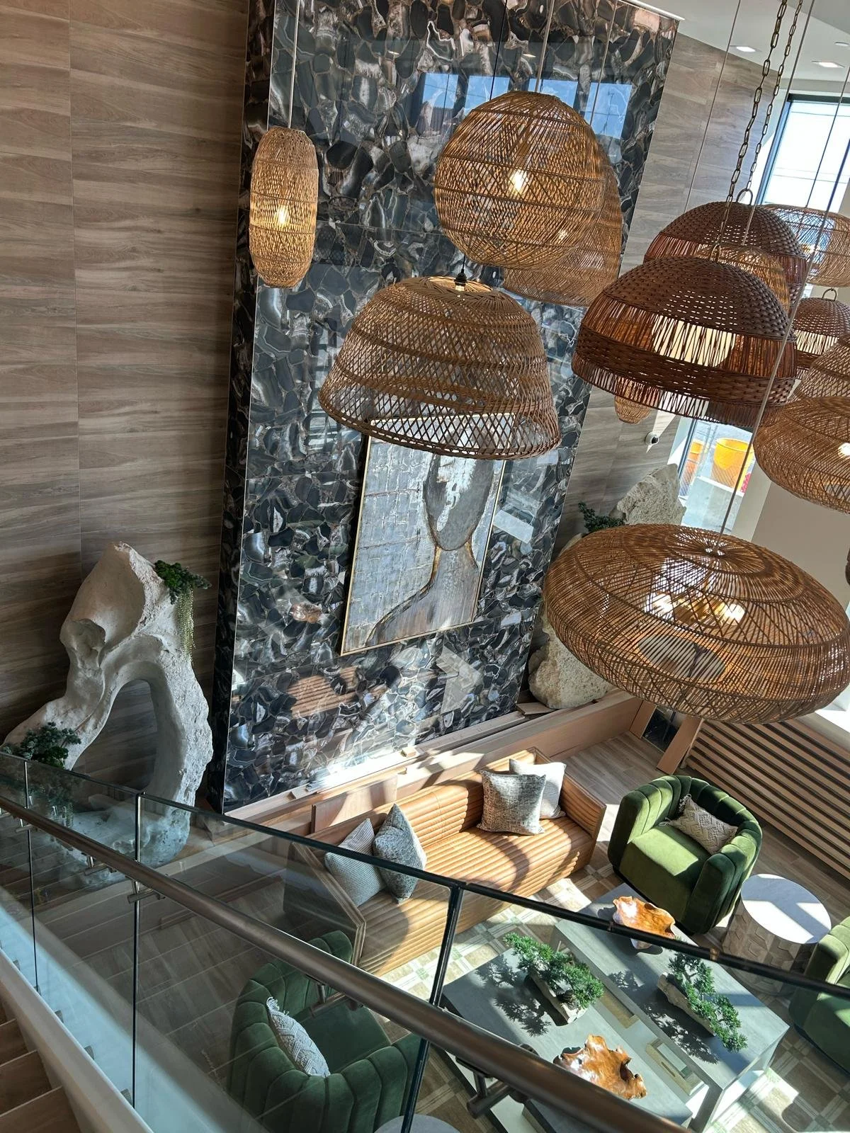 Interior view of a modern lobby or lounge area featuring large hanging wicker pendant lights, a large abstract portrait on a glossy dark stone wall, decorative rocks, and green velvet chairs around a glass coffee table with small potted plants.
