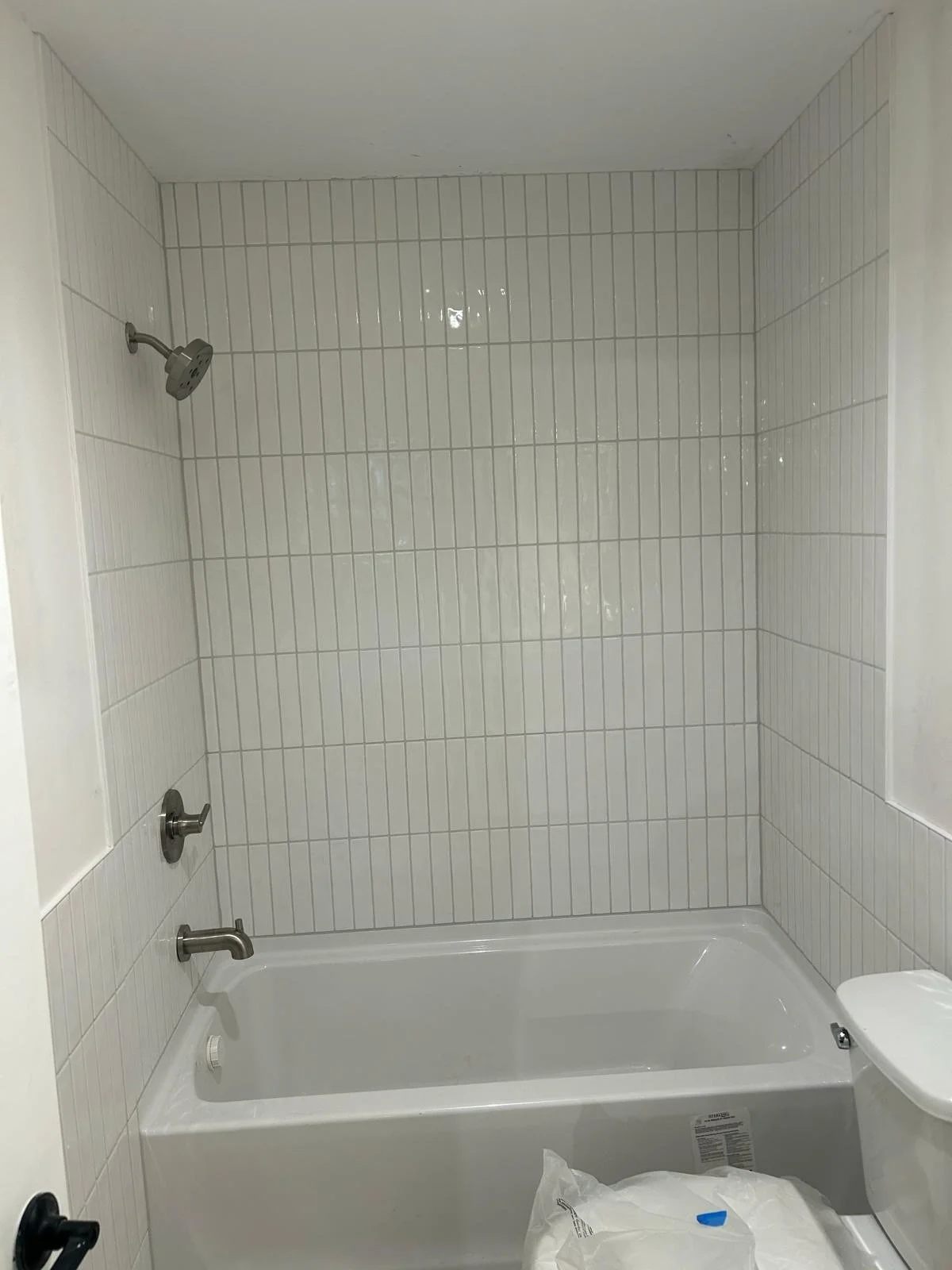Bathroom shower with white rectangular tiles, a showerhead, a bathtub, and a toilet with a plastic bag on the floor.