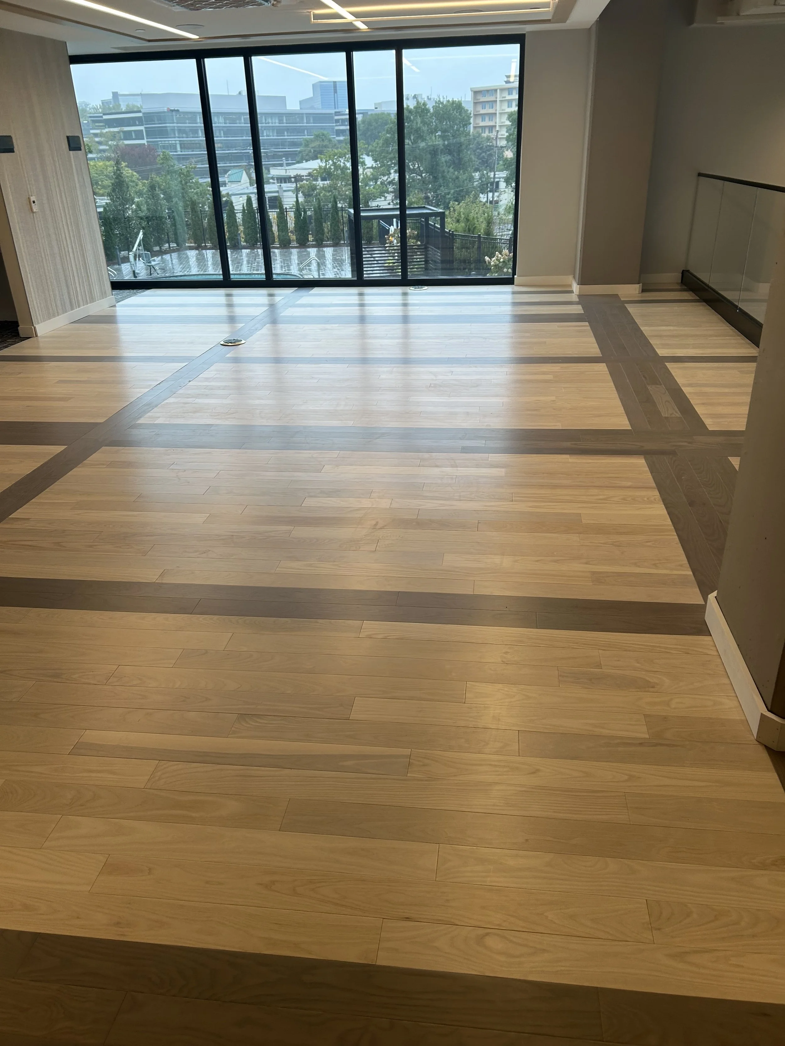 Empty room with wooden flooring and large glass windows showing an outdoor balcony and cityscape.