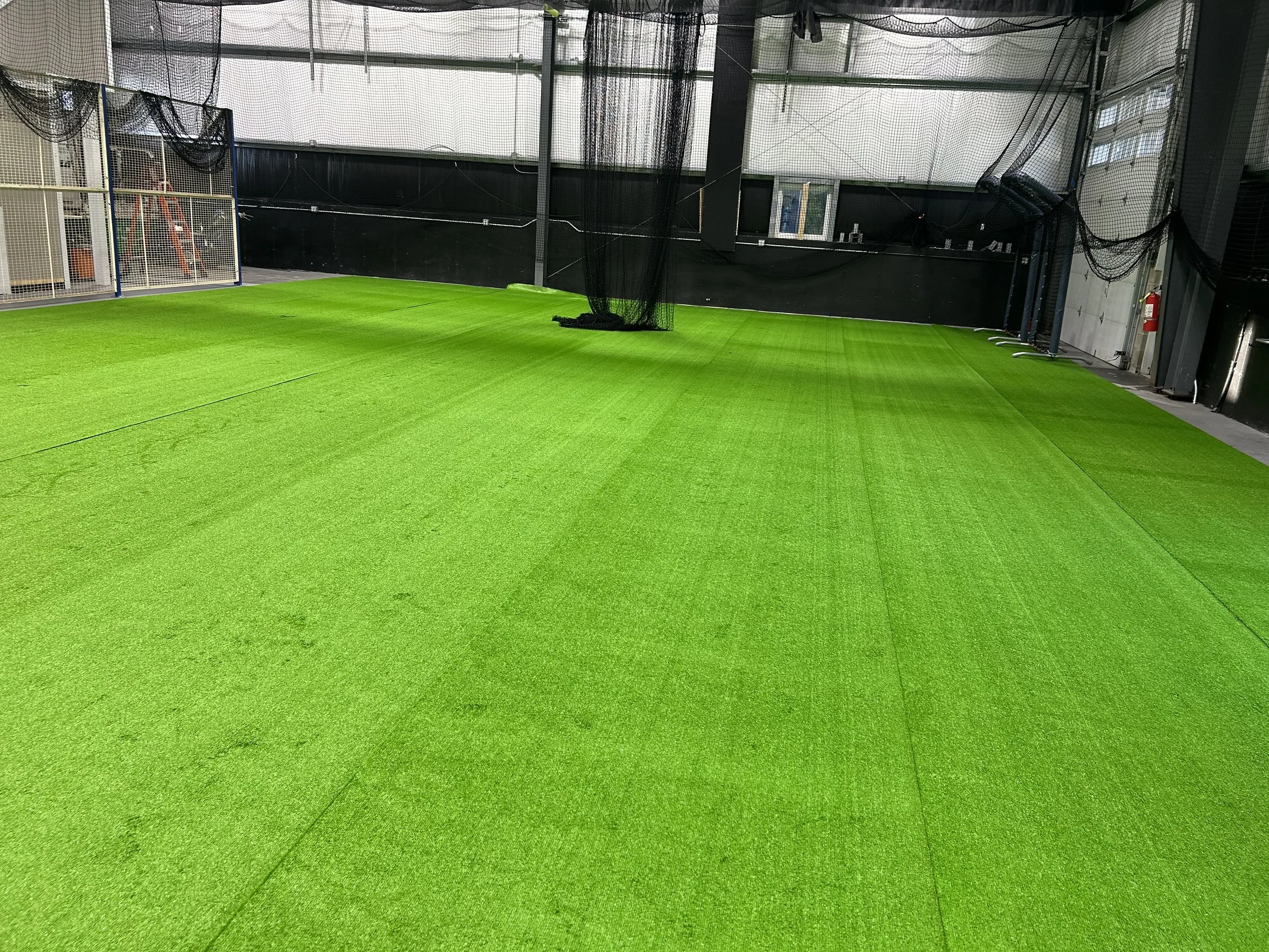 Indoor sports facility with bright green artificial turf, enclosed by black mesh netting and walls, with equipment along the sides and a fire extinguisher in the corner.