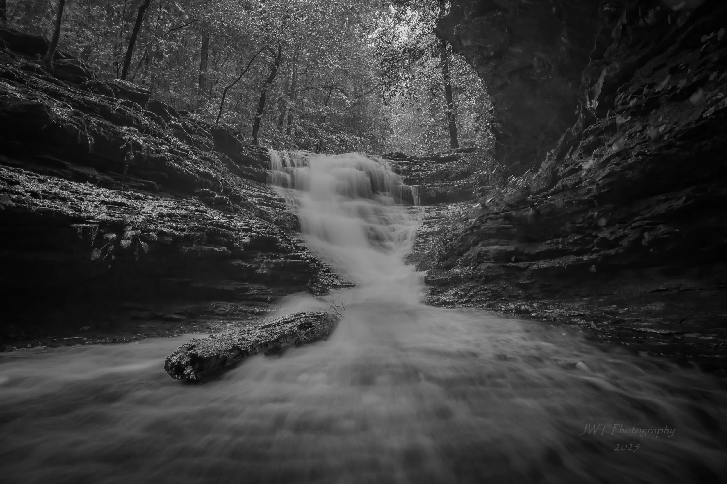 Black and white photo of a waterfall cascading over rocks in a wooded area with trees at the top and flowing water at the bottom.