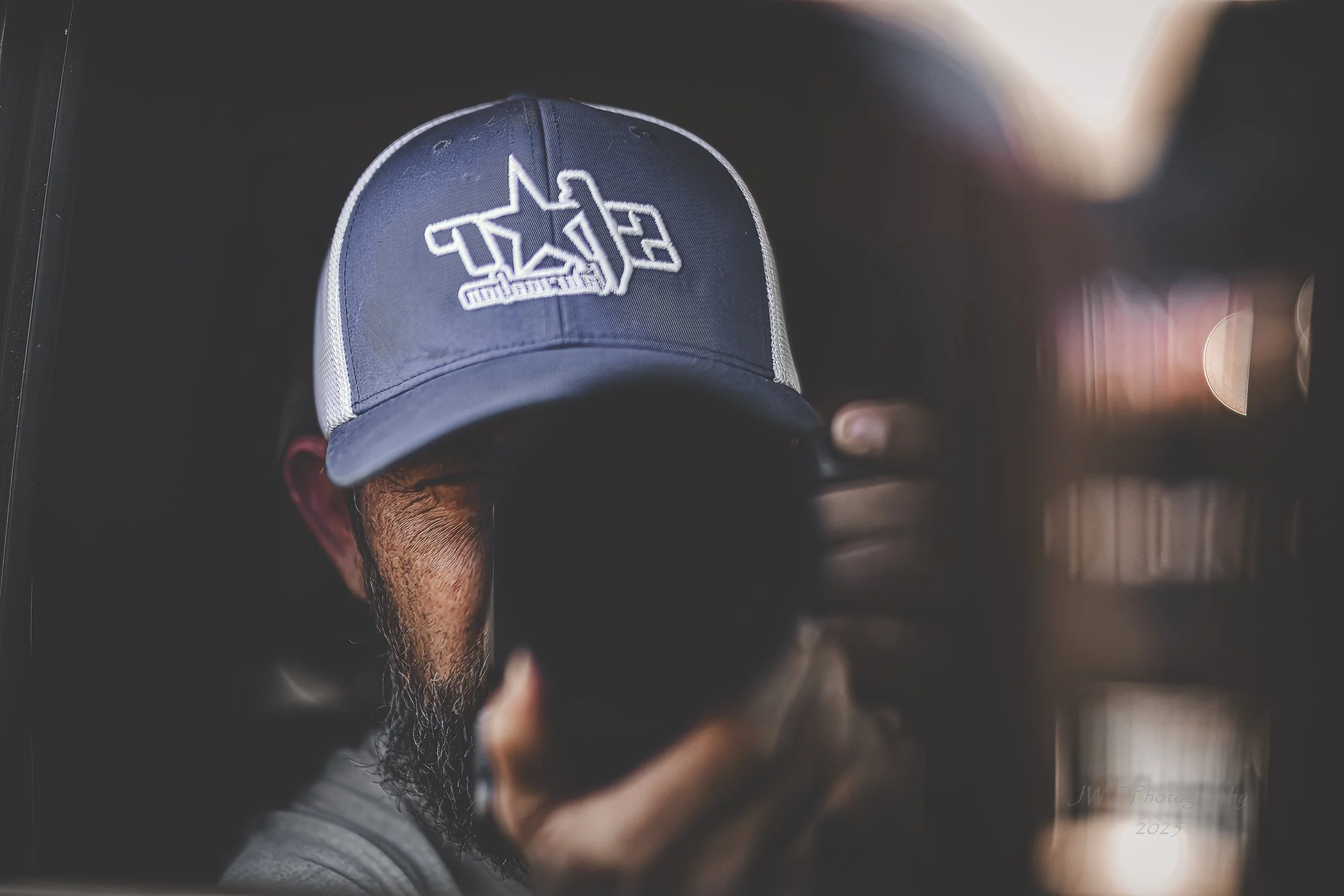 A person taking a selfie in a mirror, wearing a blue and white trucker hat with a logo. The person has a beard and is partially obscured by the black phone they are holding.
