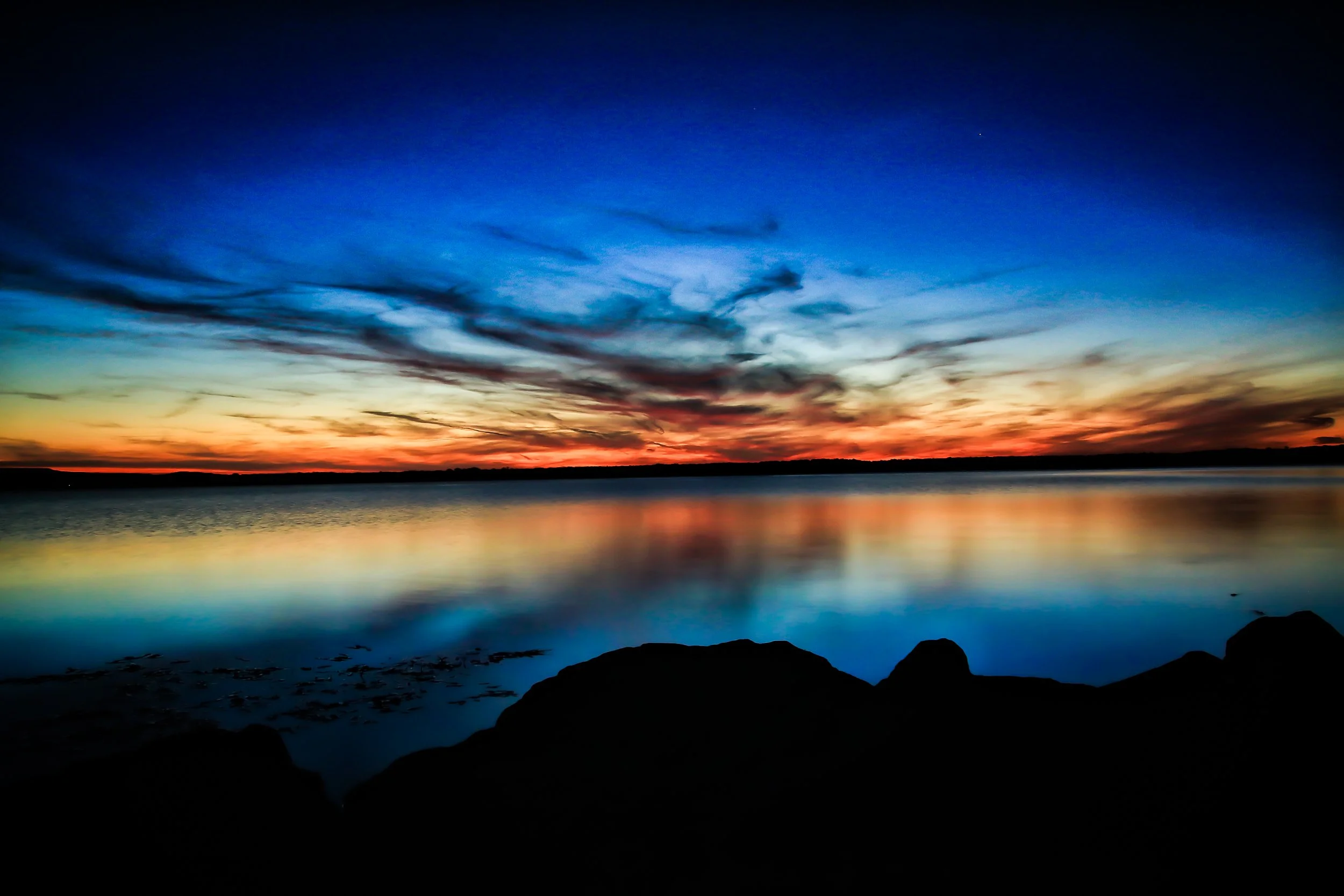 A colorful sunset over a calm body of water with rocks in the foreground and a partly cloudy sky.