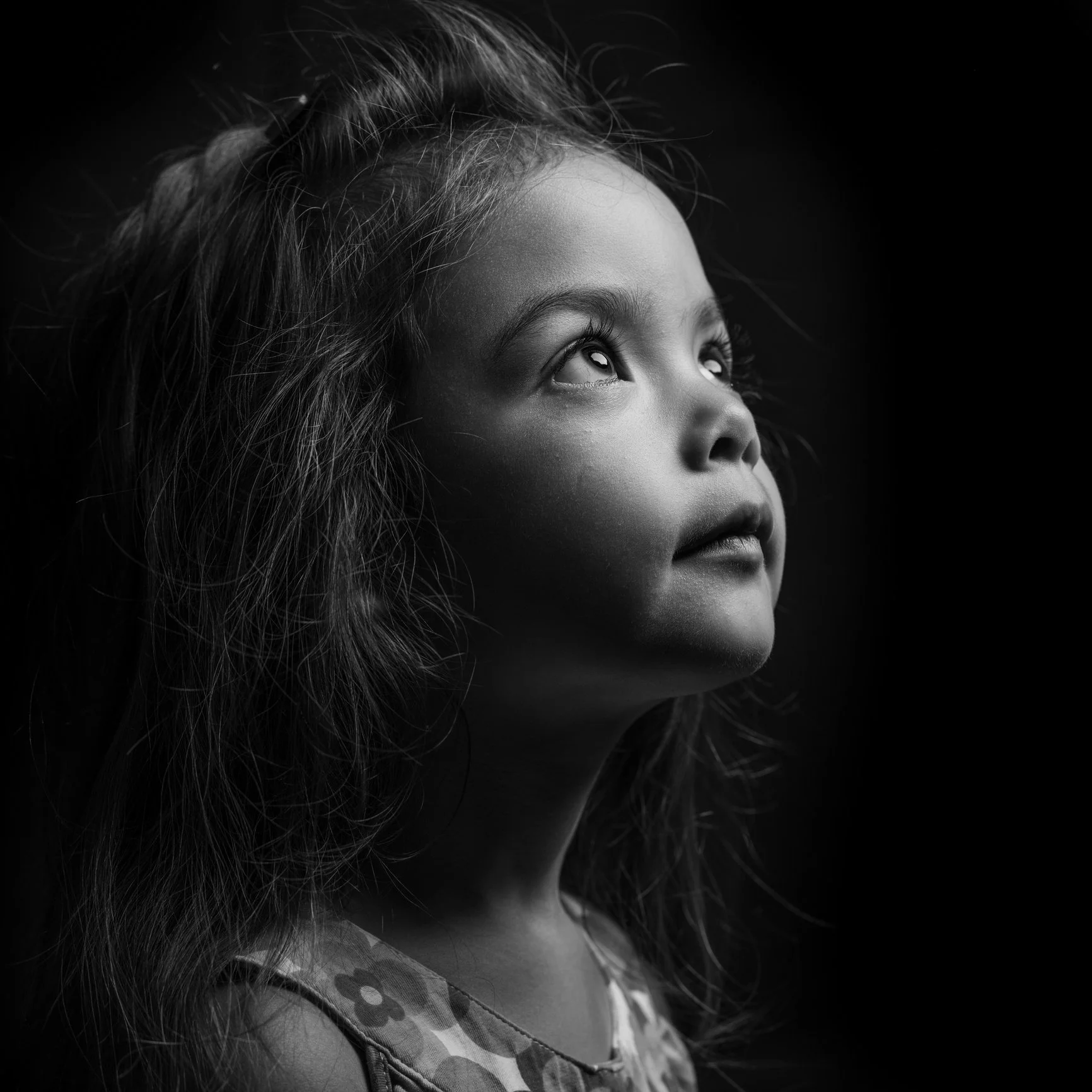 A young girl with curly hair looking upward, illuminated by soft light, in black and white.