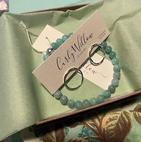 A jewelry box with a green satin lining holding a necklace and earrings with blue beads and silver rings, labeled 'Caly Millan Jewelry'.