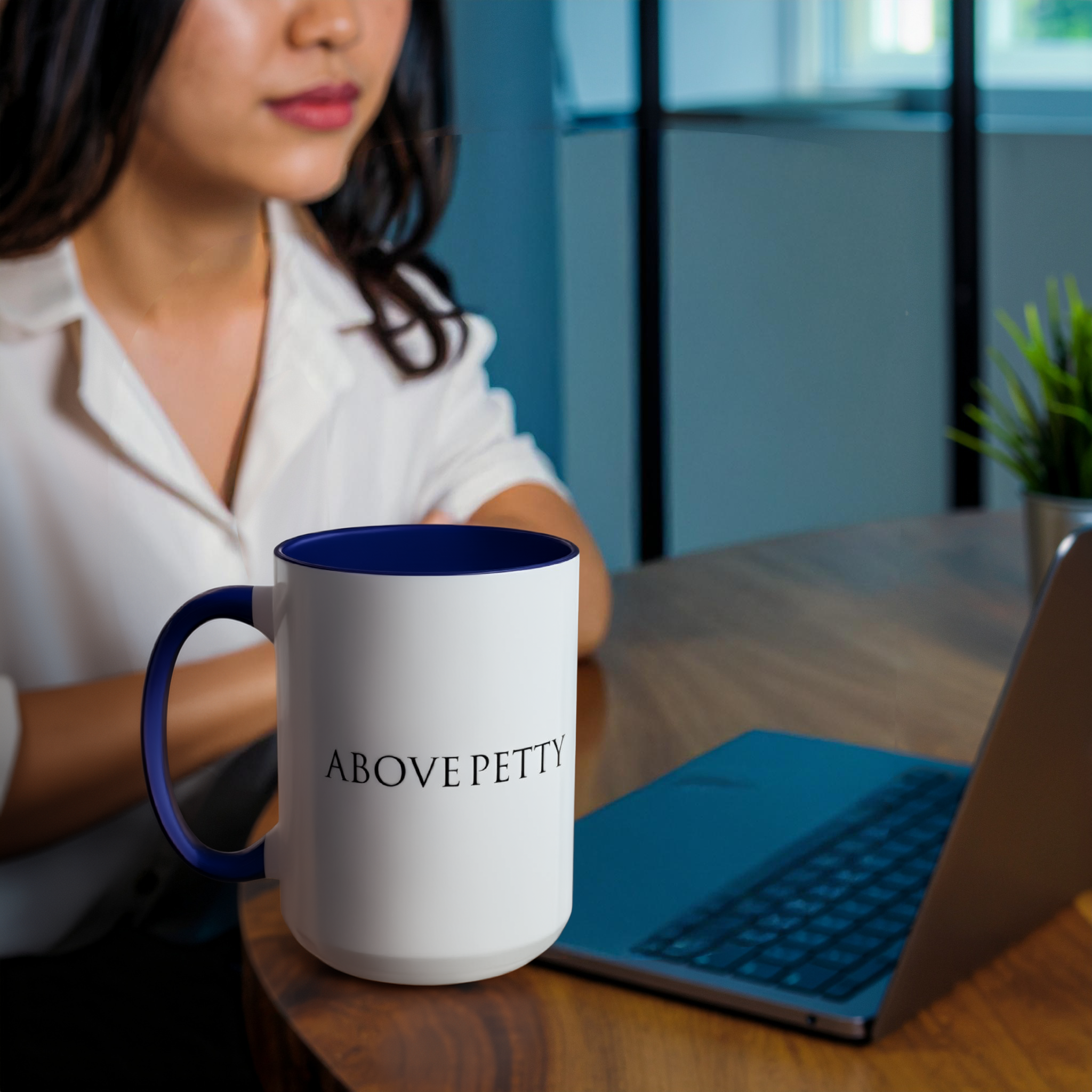 A woman sitting at a desk with a laptop and a large mug that has the words 'ABOVE PETTY' printed on it.