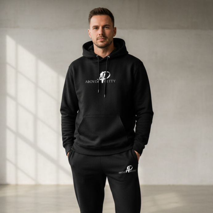 Man wearing black hoodie and sweatpants with 'Above Pretty' logo standing in a minimalist room.