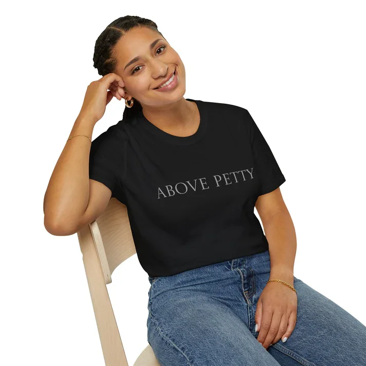 A woman sitting on a chair, smiling, wearing a black T-shirt with 'ABOVE PETTY' written on it, and blue jeans.