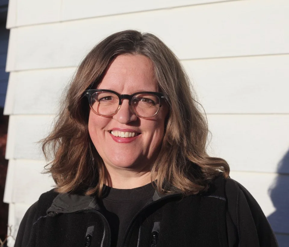 Close-up of a woman with shoulder-length brown hair, glasses, smiling, wearing a black jacket, outside with a white house siding in the background.
