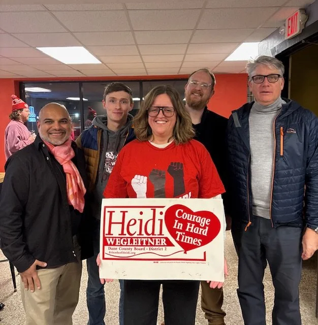 Group of five people standing indoors, holding a campaign sign for Heidi Wegleitner, with a woman in the middle wearing a red shirt with raised fists graphic and the campaign slogan 'Courage In Hard Times'.