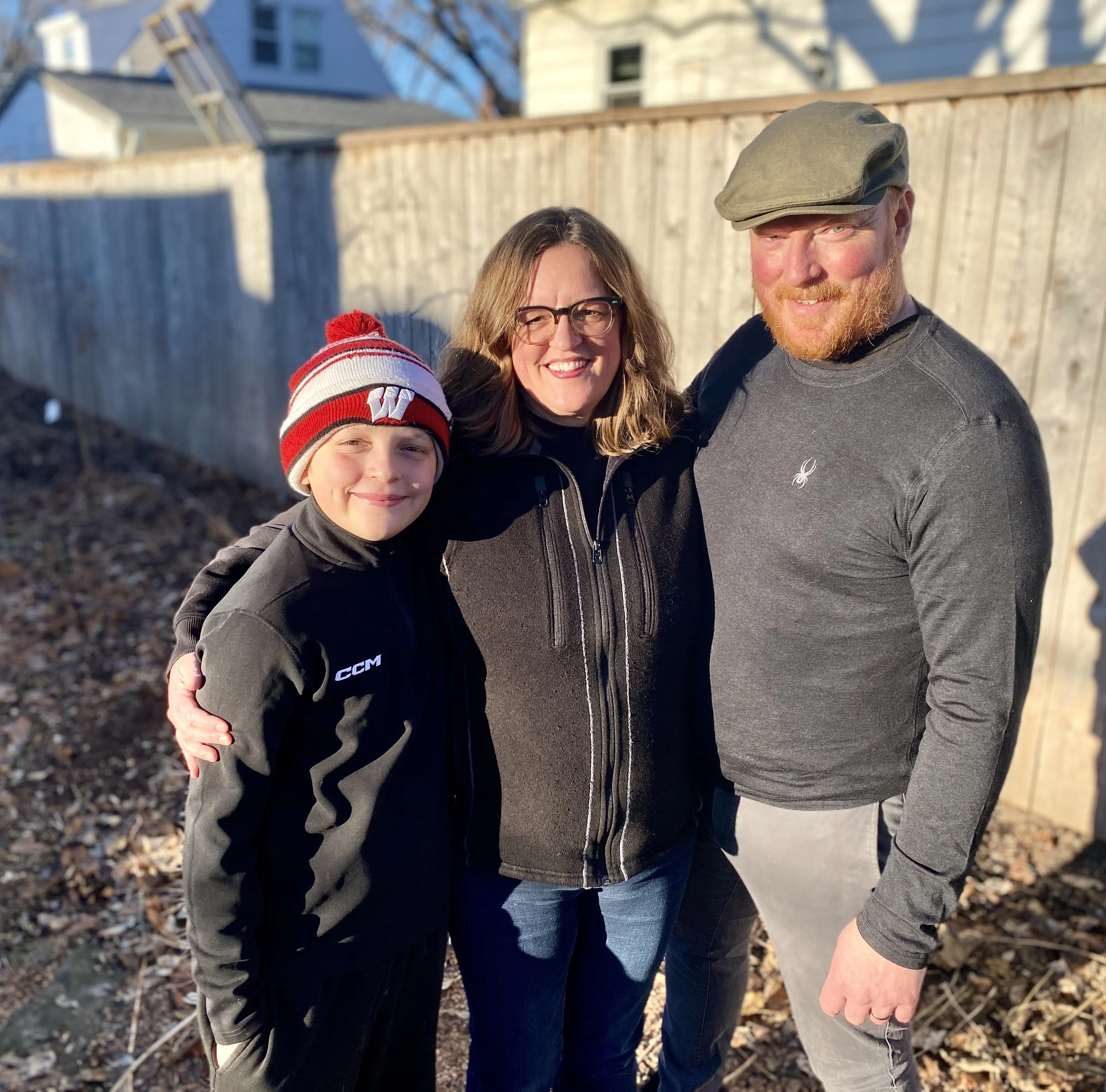 A woman, a boy, and a man stand outdoors in front of a wooden fence, smiling and embracing each other on a sunny day.