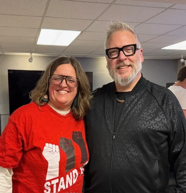 Two smiling people, a woman and a man, standing indoors with ceiling lights, both wearing glasses. The woman is wearing a red T-shirt with protesting fists and the words "STAND" on it. The man is wearing a black jacket.