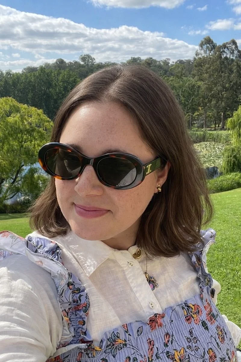 A woman with shoulder-length brown hair wearing black sunglasses, earrings, a necklace, and a white blouse with floral ruffles, outdoors in a green park with trees and water in the background under a partly cloudy sky.