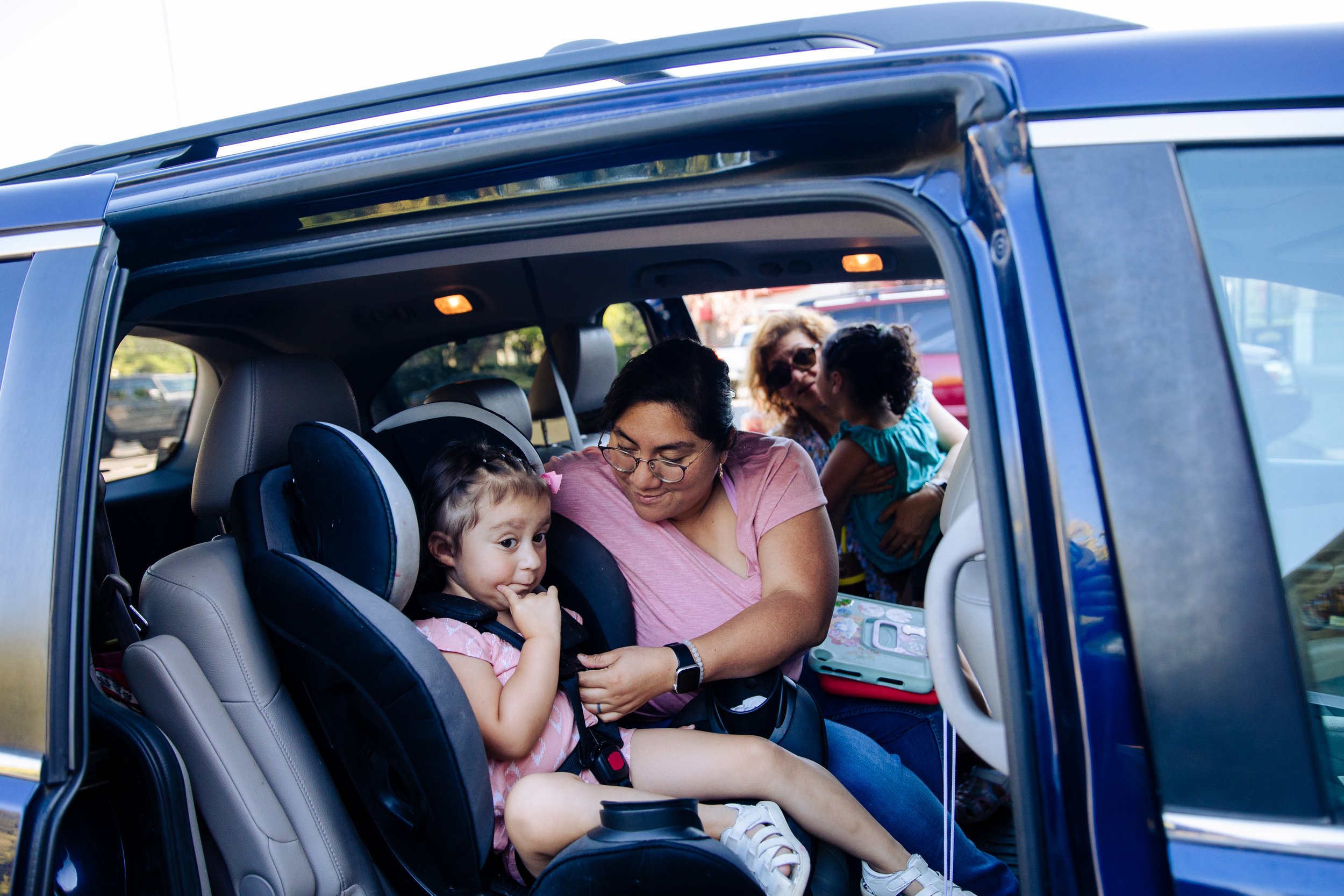 A woman and two children sitting in a car with one child in a booster seat. They are in a parking lot, with others parked in the background. The woman is looking at the child in the booster seat, while another woman and a child are outside the car looking inside.