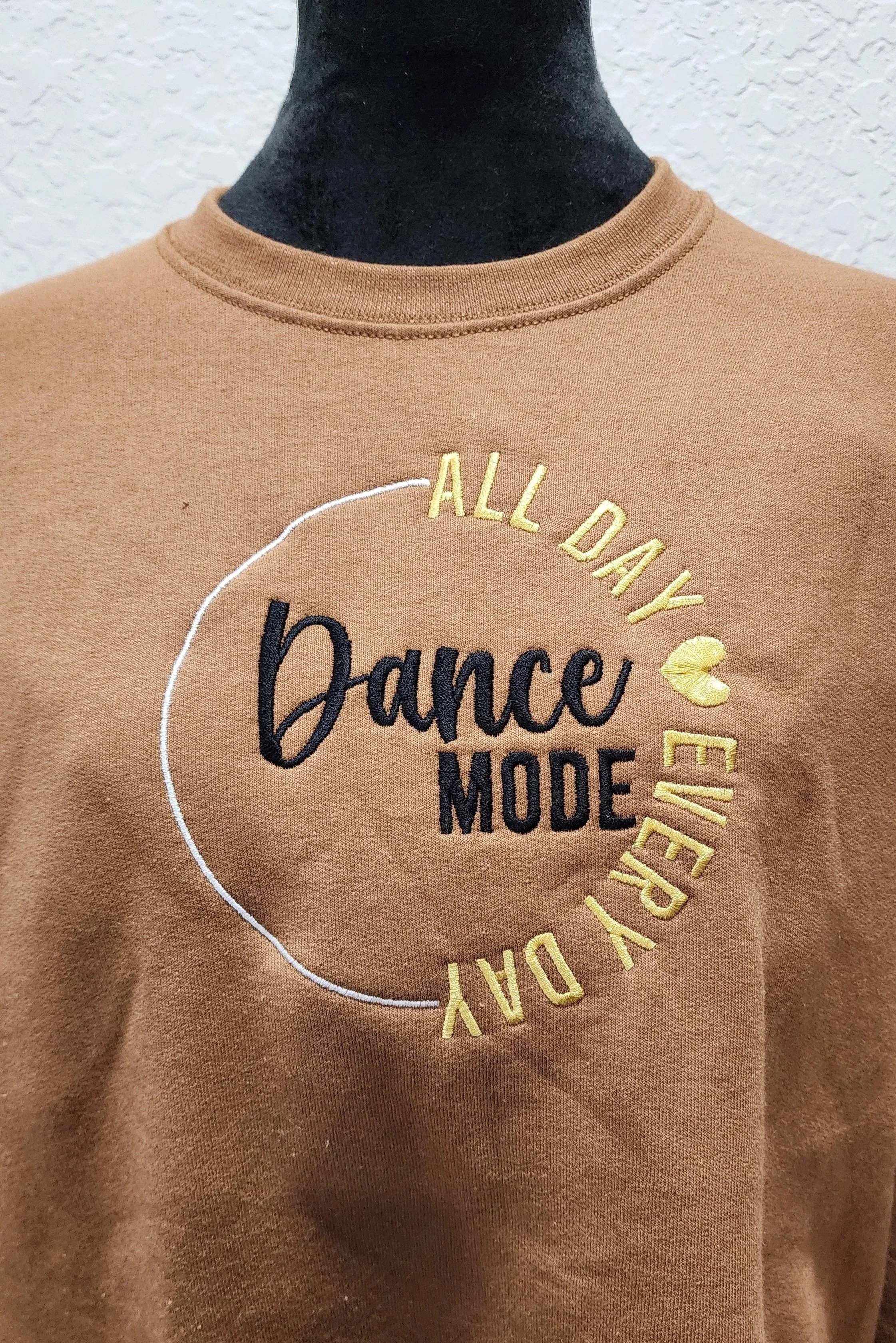 light brown sweatshirt on a mannequin with embroidered design in black, gold and white. the design says dance mode all day every day