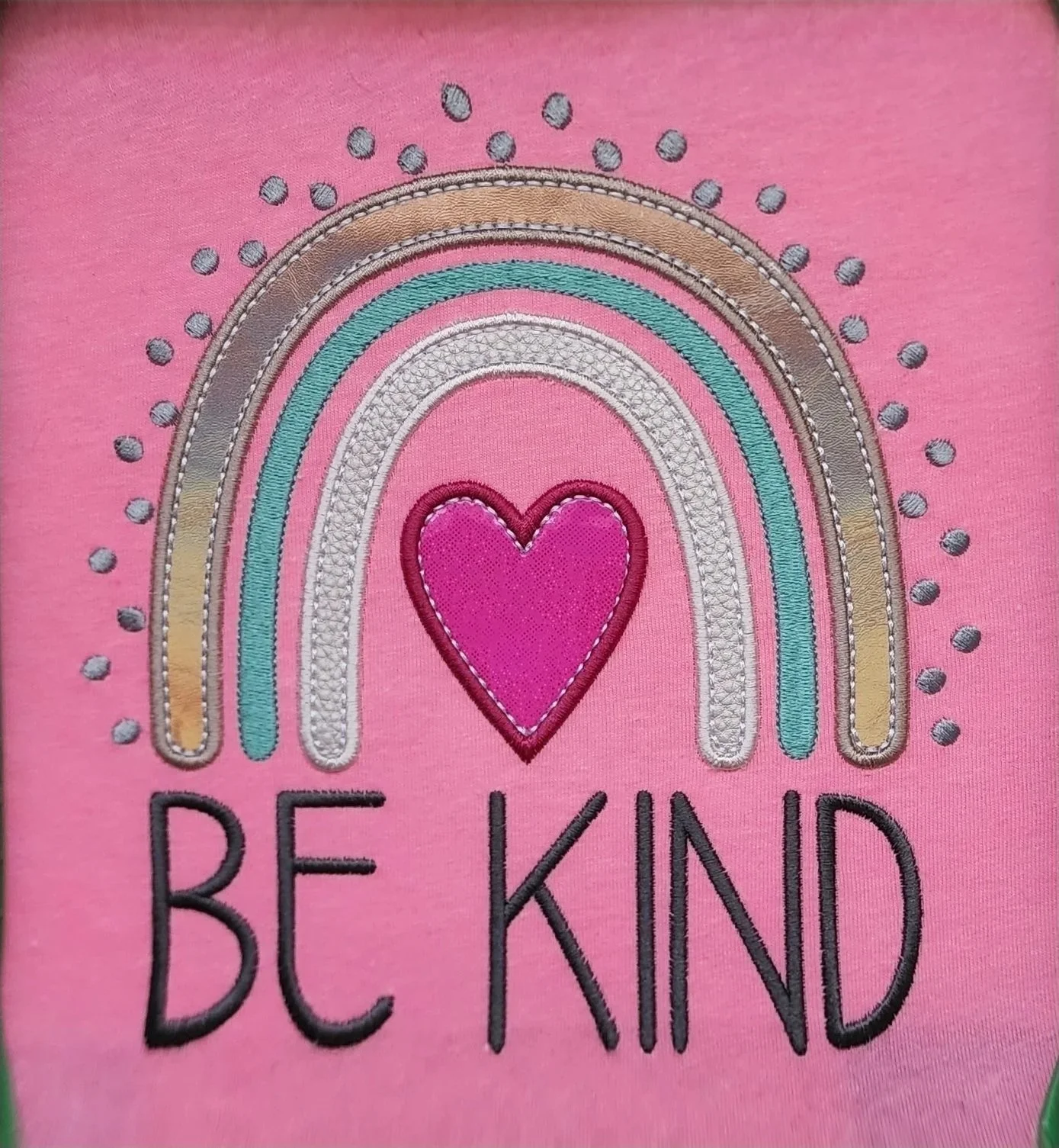 applique of a rainbow and a heart and embroidered text "be kind" on a pink fabric