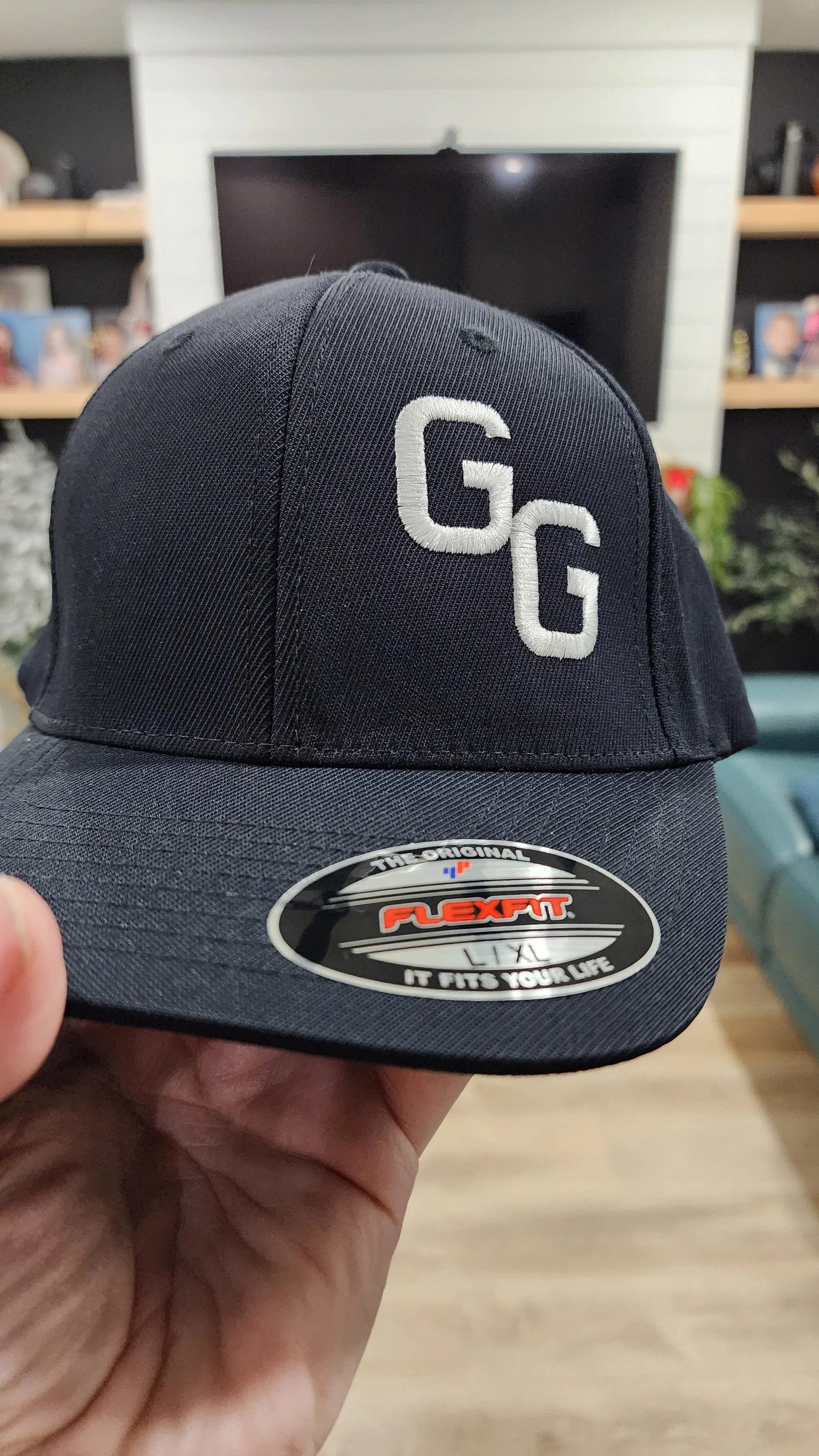 A black baseball cap with white embroidered 'GG' initials on the front, held in a person's hand indoors in front of a television and shelves with photos and decorations.