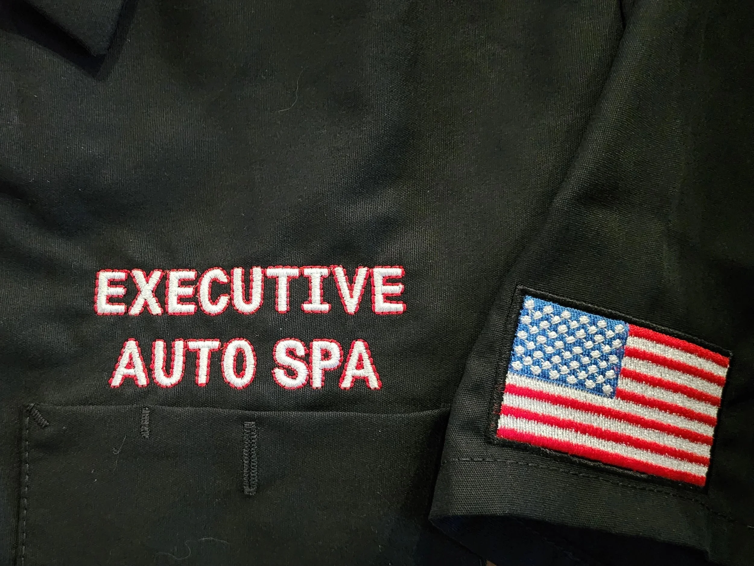 Black shirt with close-up embroidered logo in white lettering and red outline, embroidered flag in a sleeve
