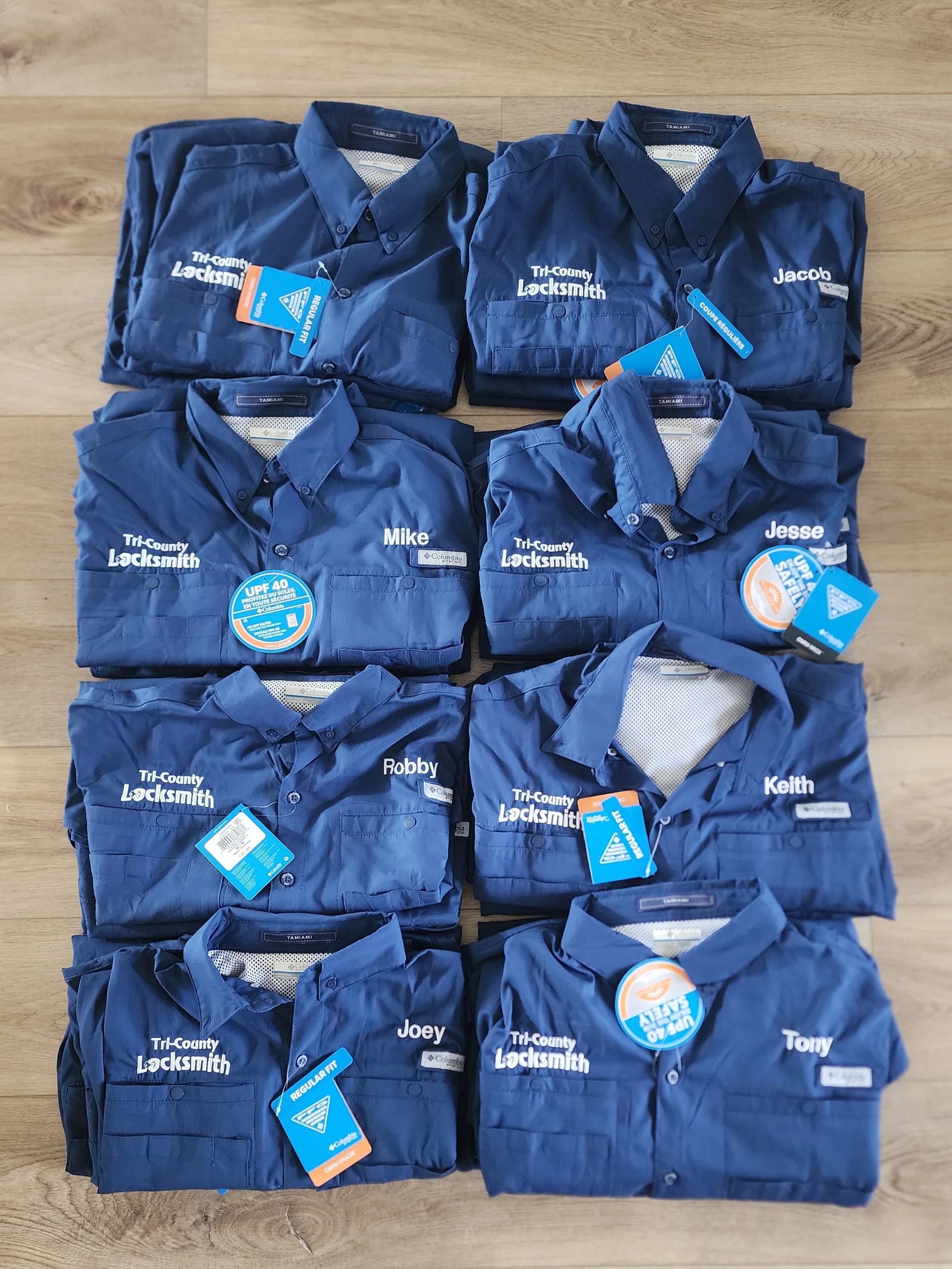 Nine blue work shirts, each embroidered with names and 'Tri-County Locksmith,' laid out on a wooden surface.