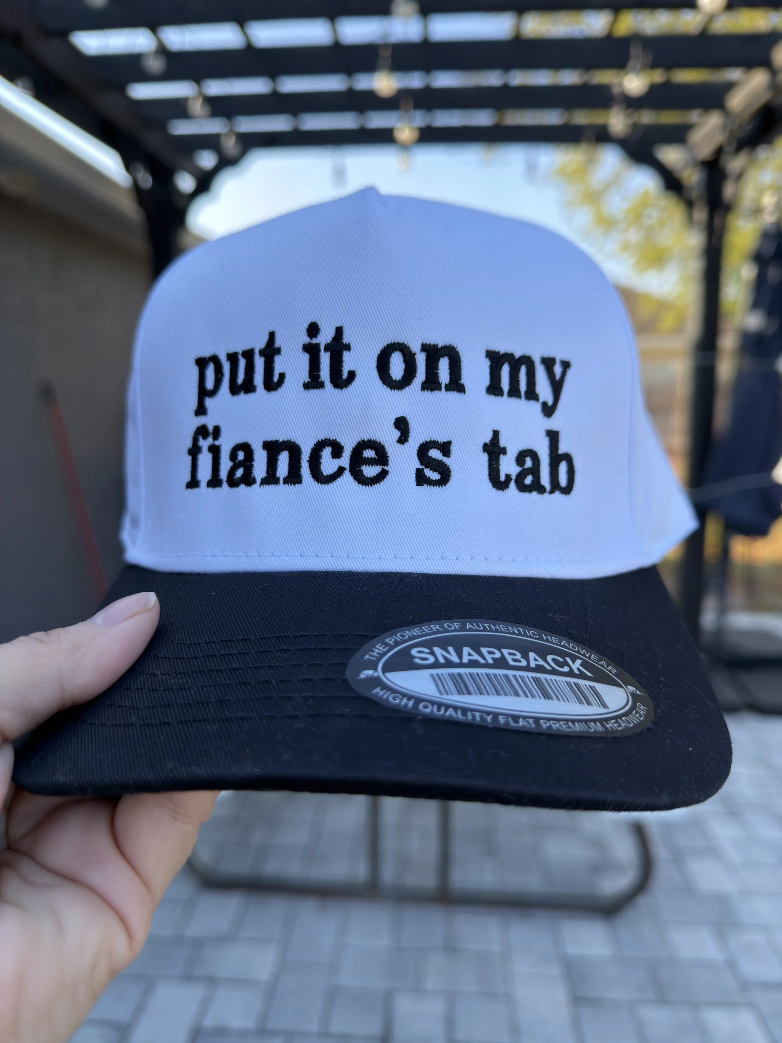 white and black trucker hat with embroidered "put it on my fiance's tab"
