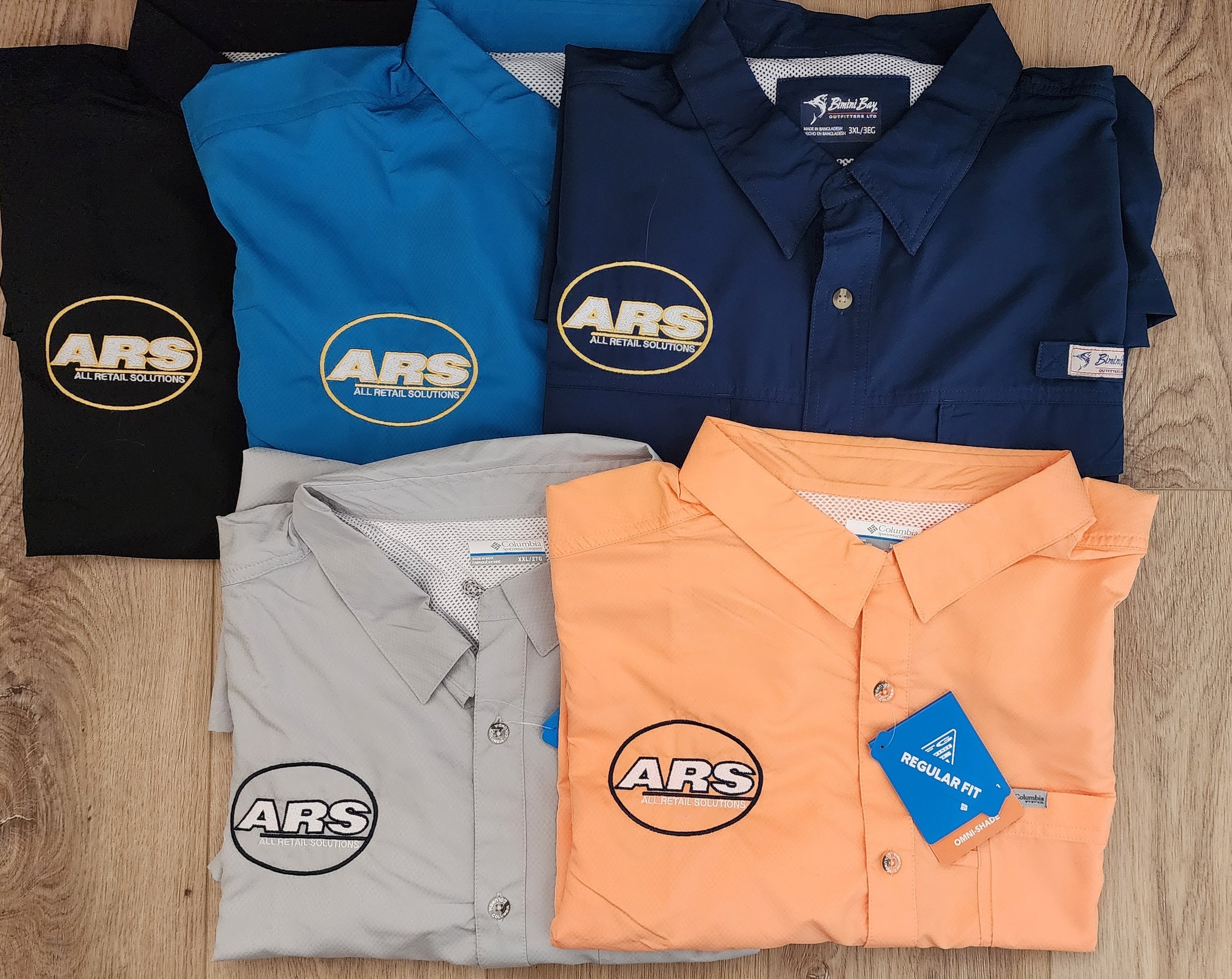 Five collared shirts in black, blue, dark blue, light gray, and peach, each with a logo that reads 'ARS All Retail Solutions'. The peach shirt has a blue tag indicating a regular fit. The shirts are neatly folded and arranged on a wood surface.