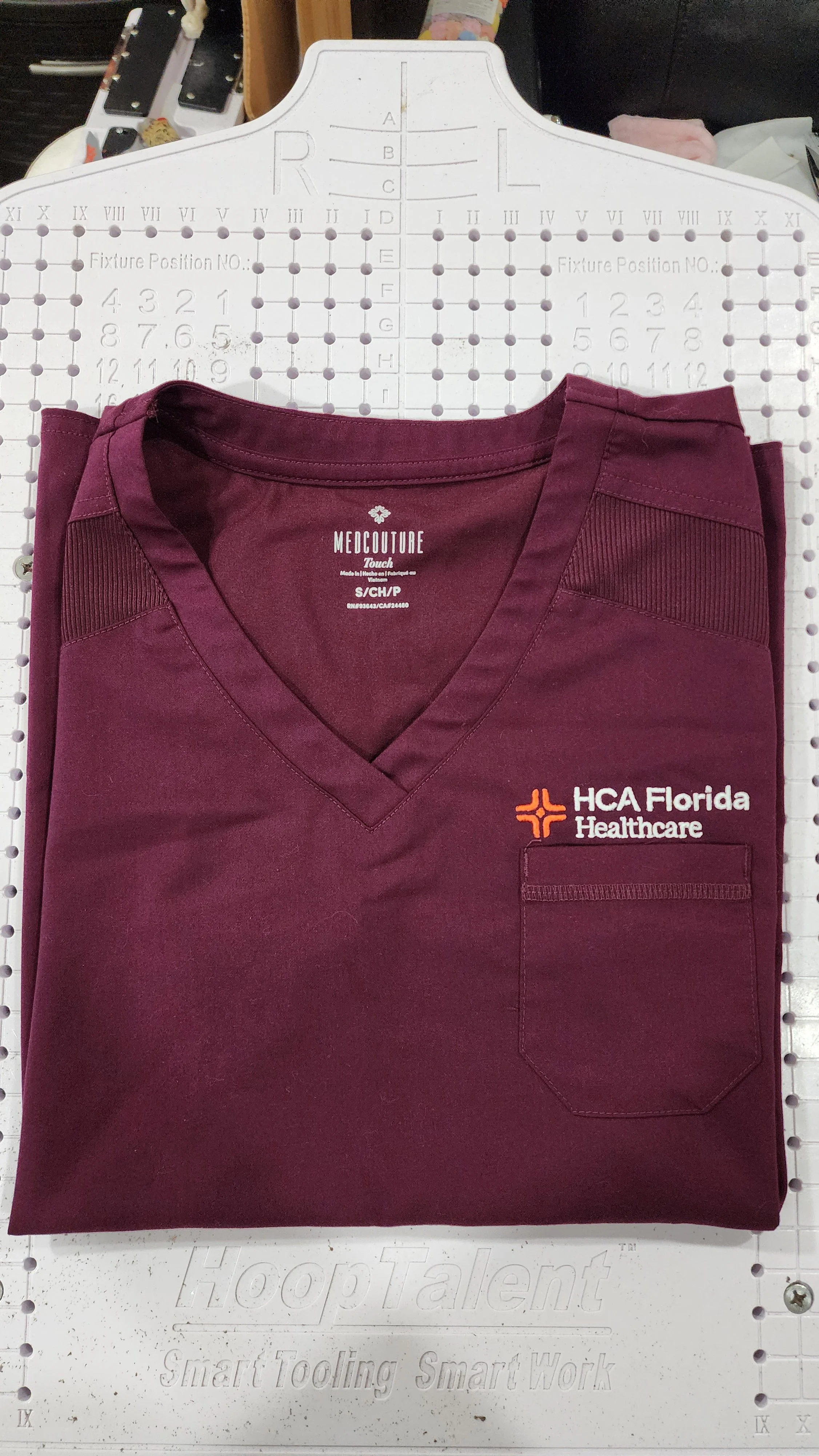 A folded maroon medical scrub top with the HCA Florida Healthcare logo and text embroidered on the front pocket.