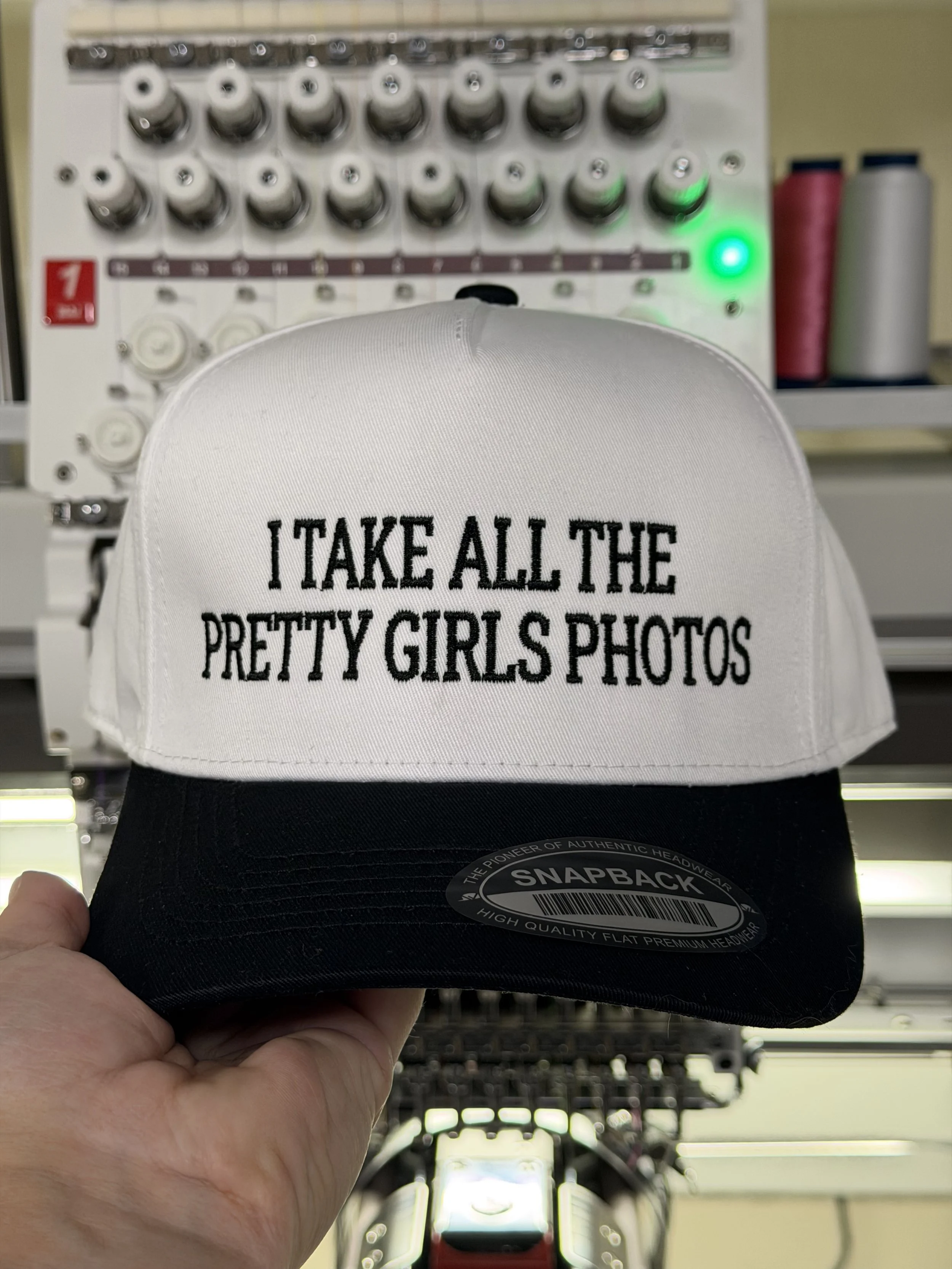 hand holding a black and white trucker hat with embroidered "I take all the pretty girls photos"