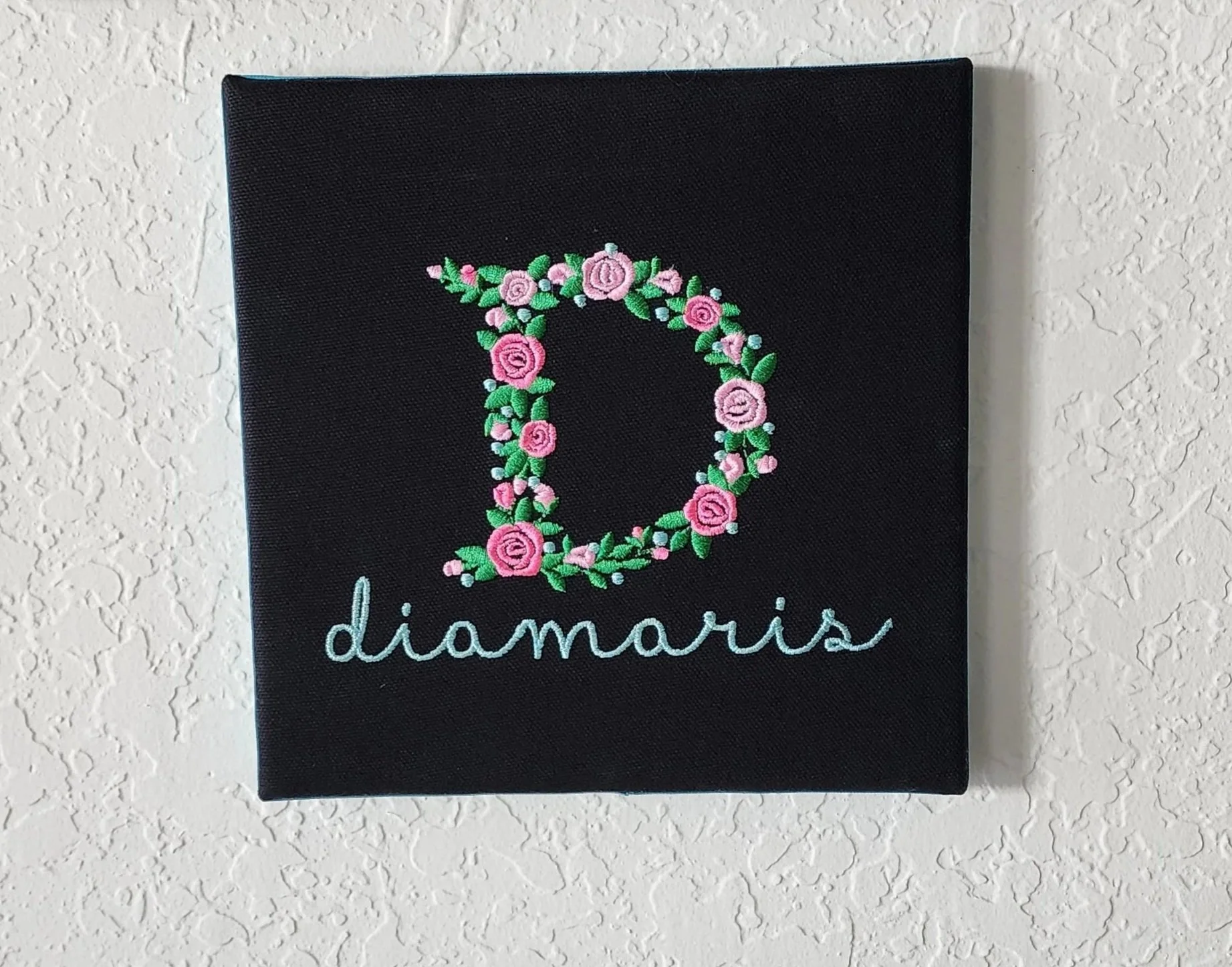 black square canvas picture with embroidered flower initial and a name underneath