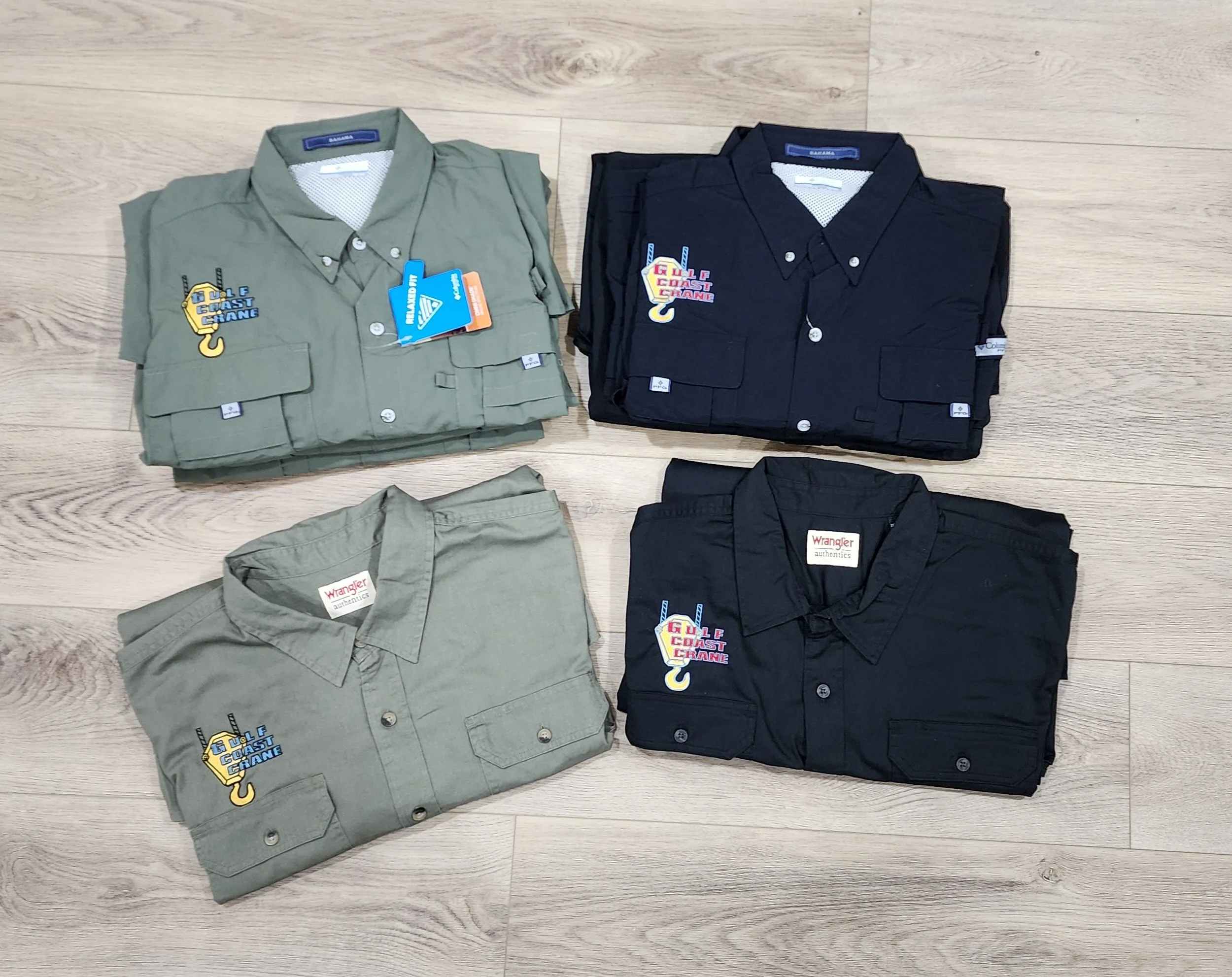 Four folded work shirts on a light wood floor, two in green and two in black, with logos and tags attached.