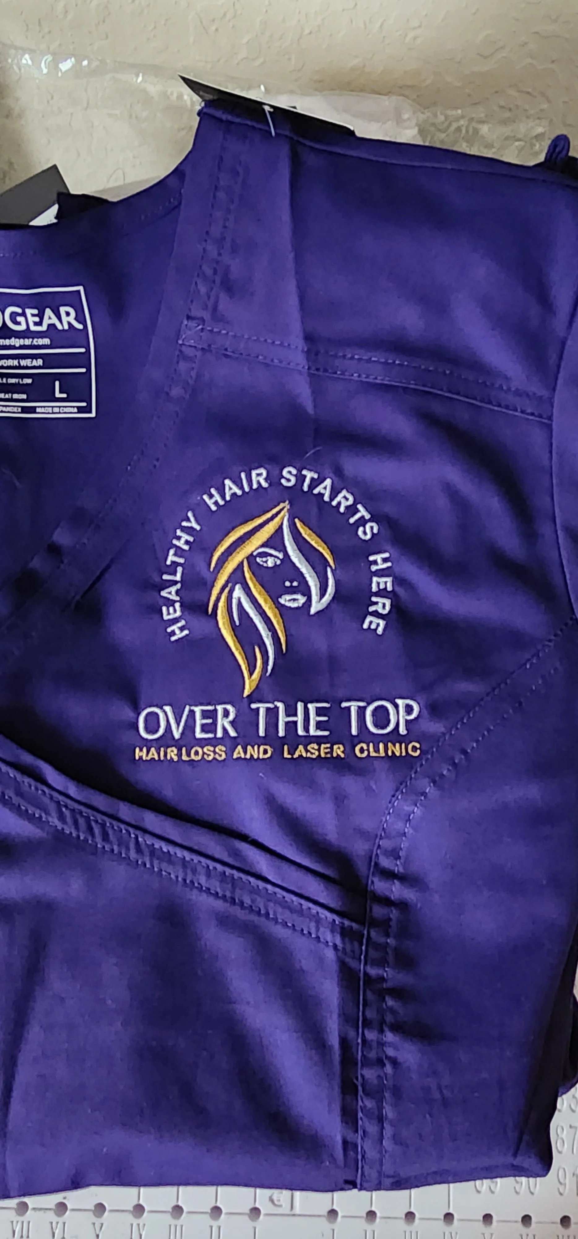 A purple garment, likely scrubs, with an embroidered logo of a woman's face with hair and the text 'Healthy Hair Starts Here Over The Top Hair Loss and Laser Clinic'.
