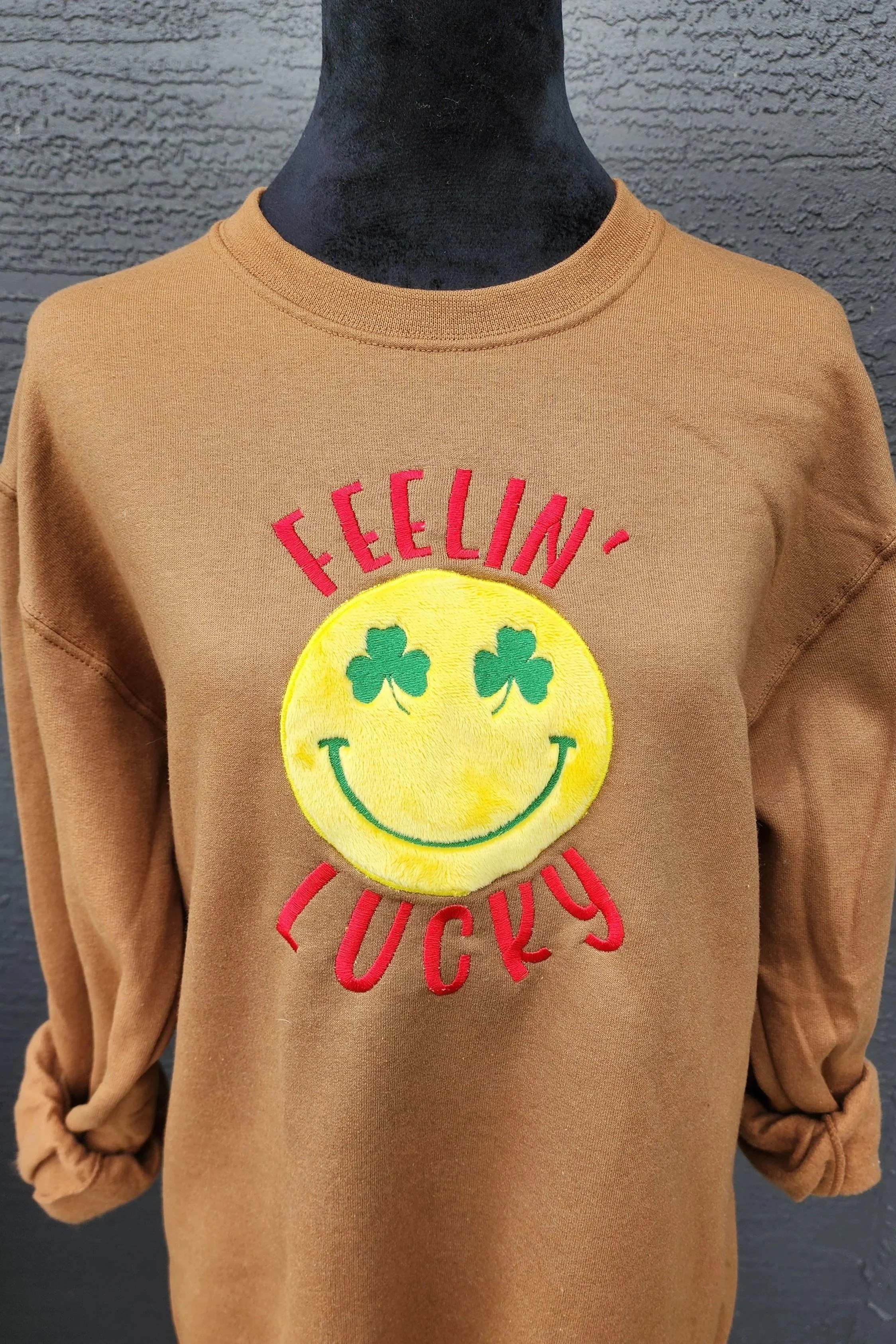 Light brown sweatshirt on a mannequin with embroidered design. the design is an applique of a yellow emoji with green clover as eyes and a smile. embroidered feelin' lucky