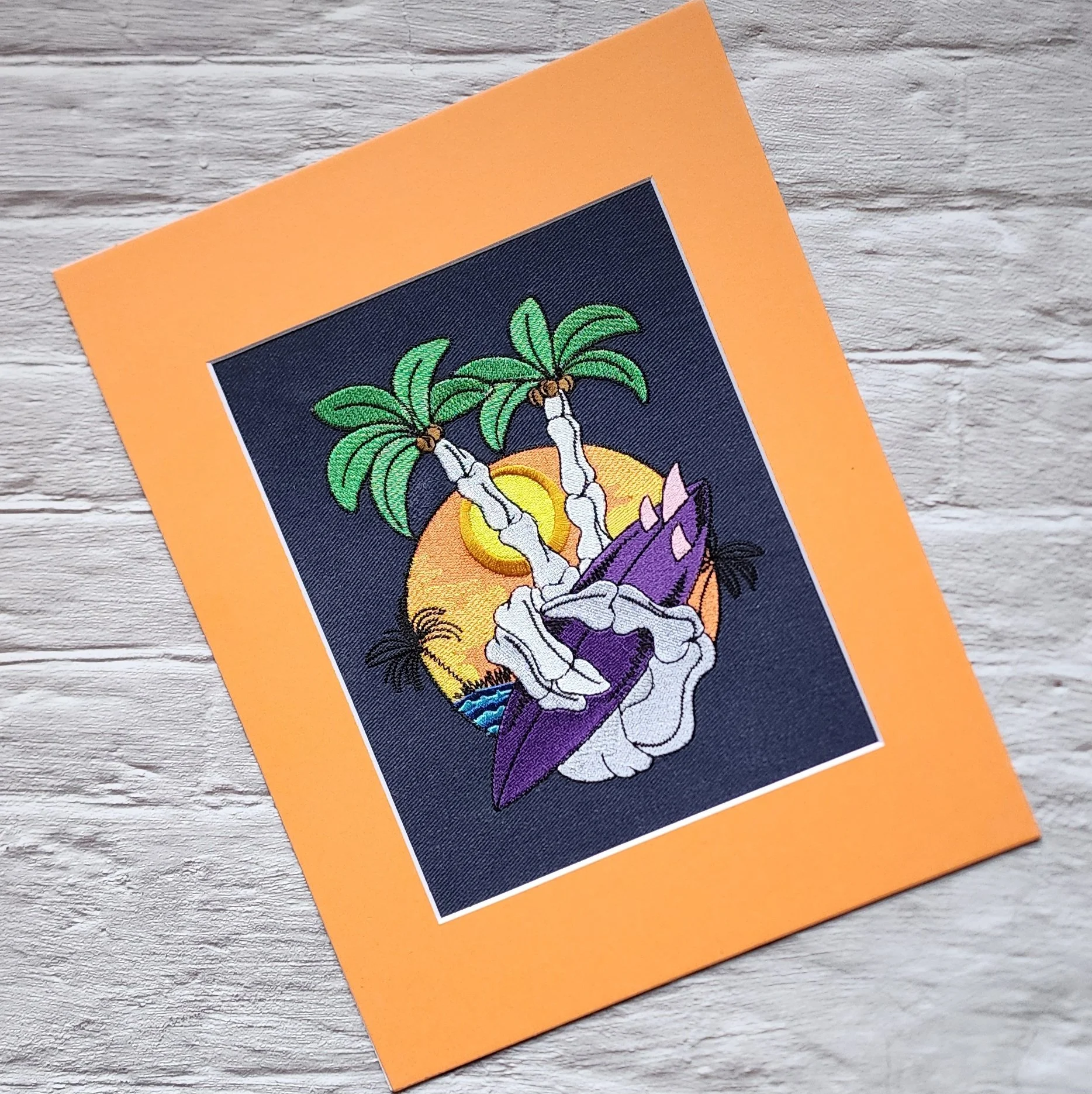 matted wall art with embroidered design of a skeleton hand holding a surf board and making a peace sign with pointer and middle fingers sticking out and look like palm trees, sunset in the background