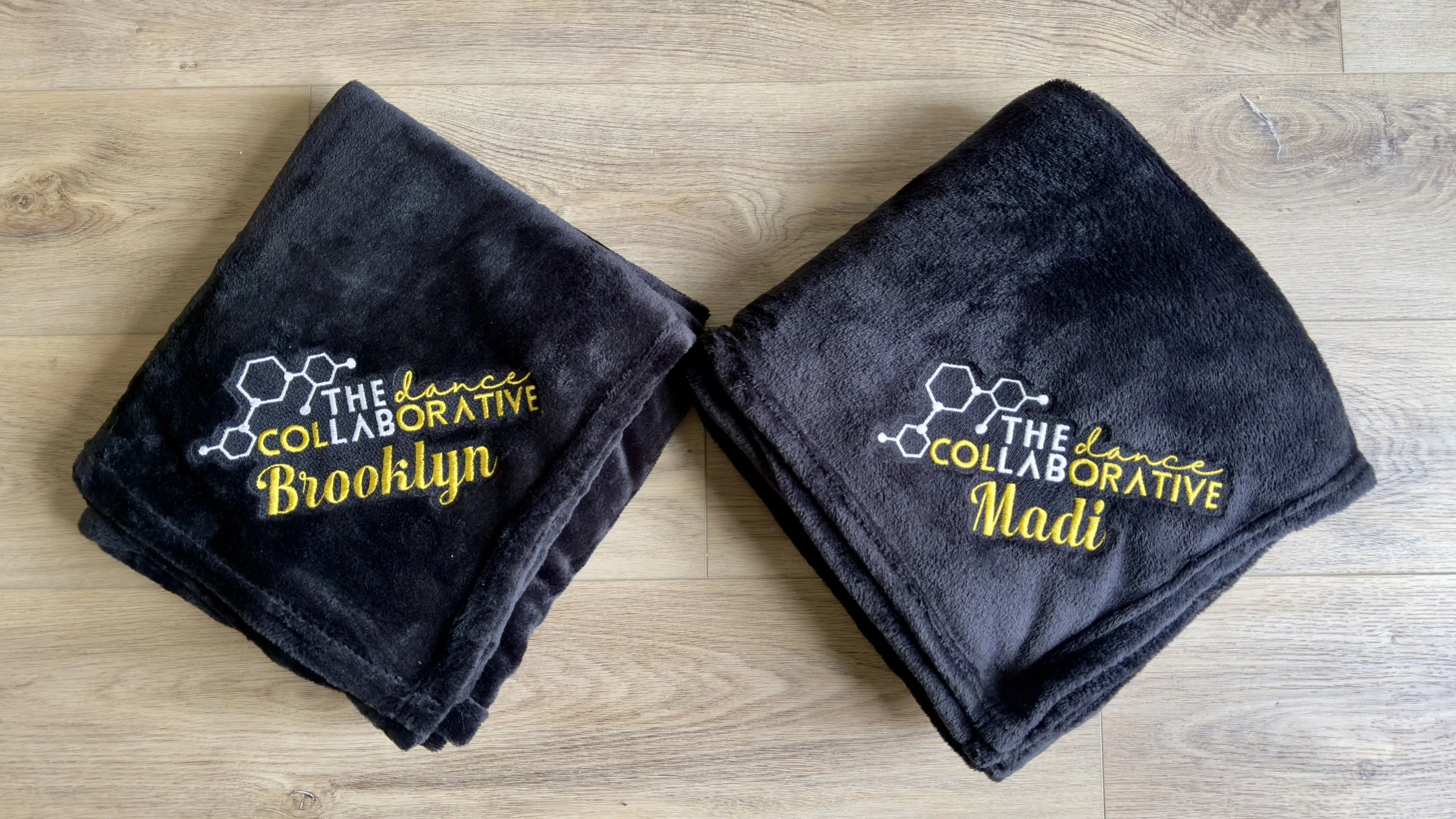 two black plush blankets with a business logo and a name embroidered in a corner, displayed on a wooden background