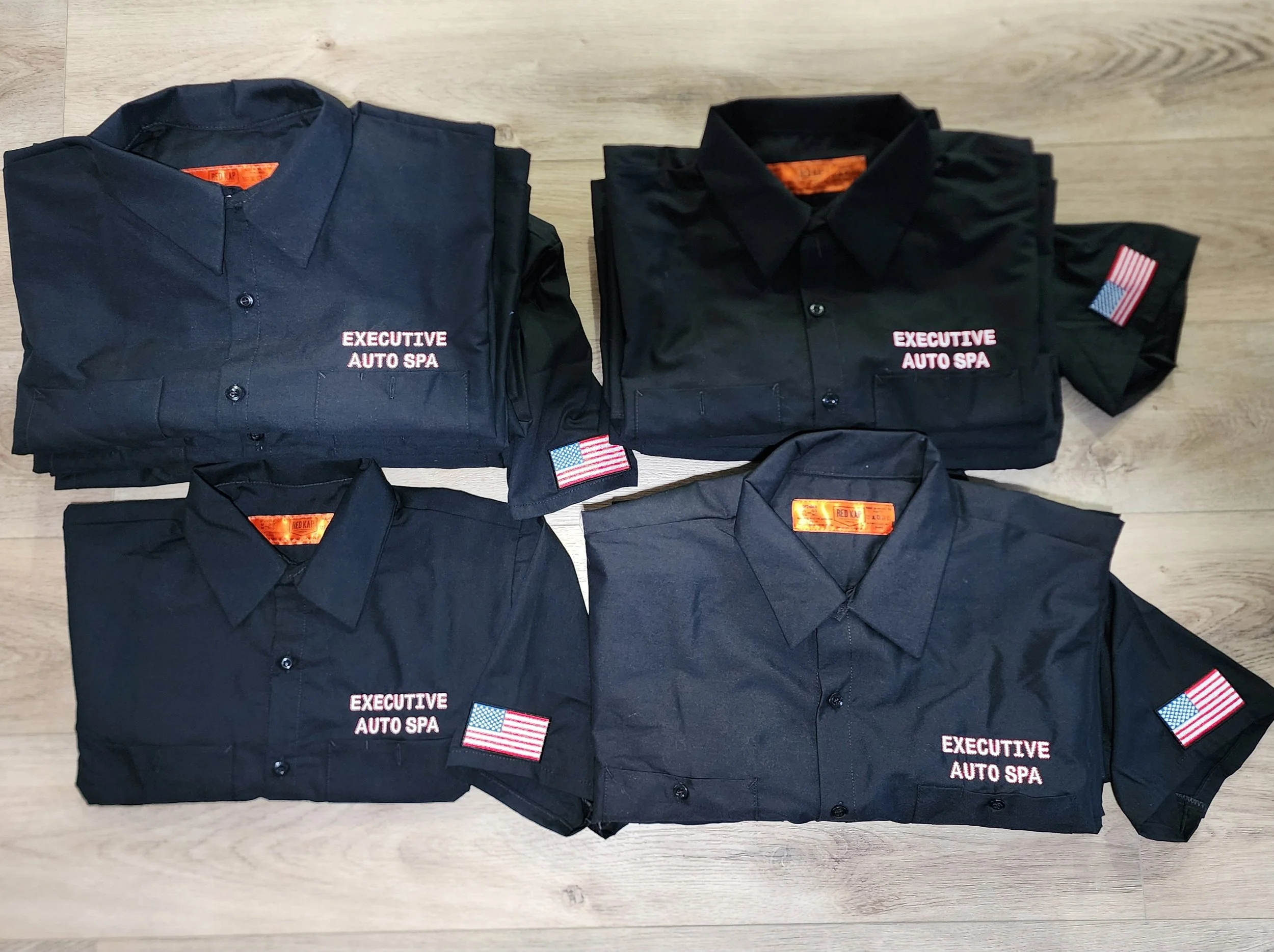 Four dark blue uniforms with patches that read "EXECUTIVE AUTO SPA" and American flag patches, folded and laid out on a wooden floor.