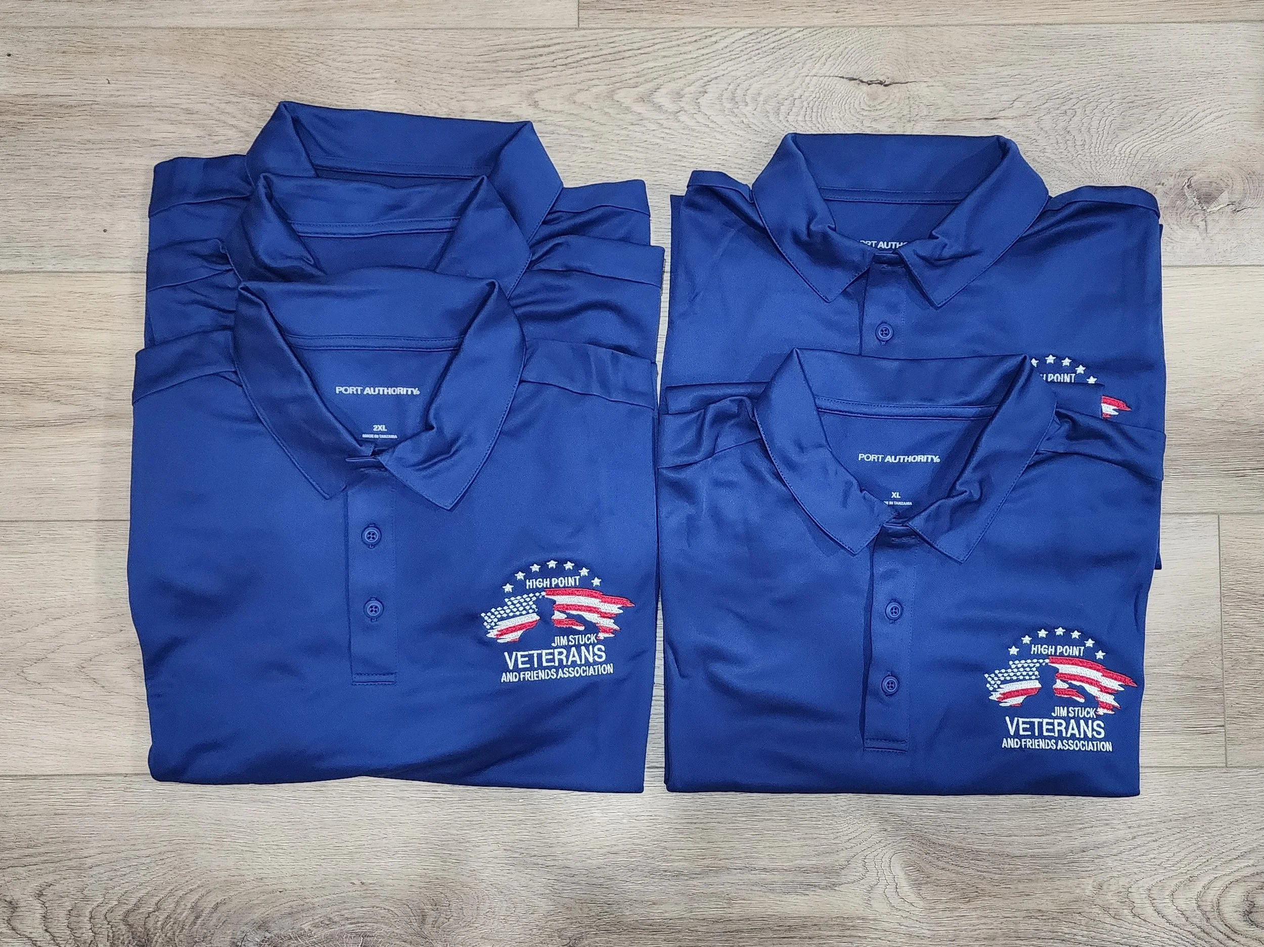 Six folded blue polo shirts with embroidered logos and text, laid out on a wooden floor.