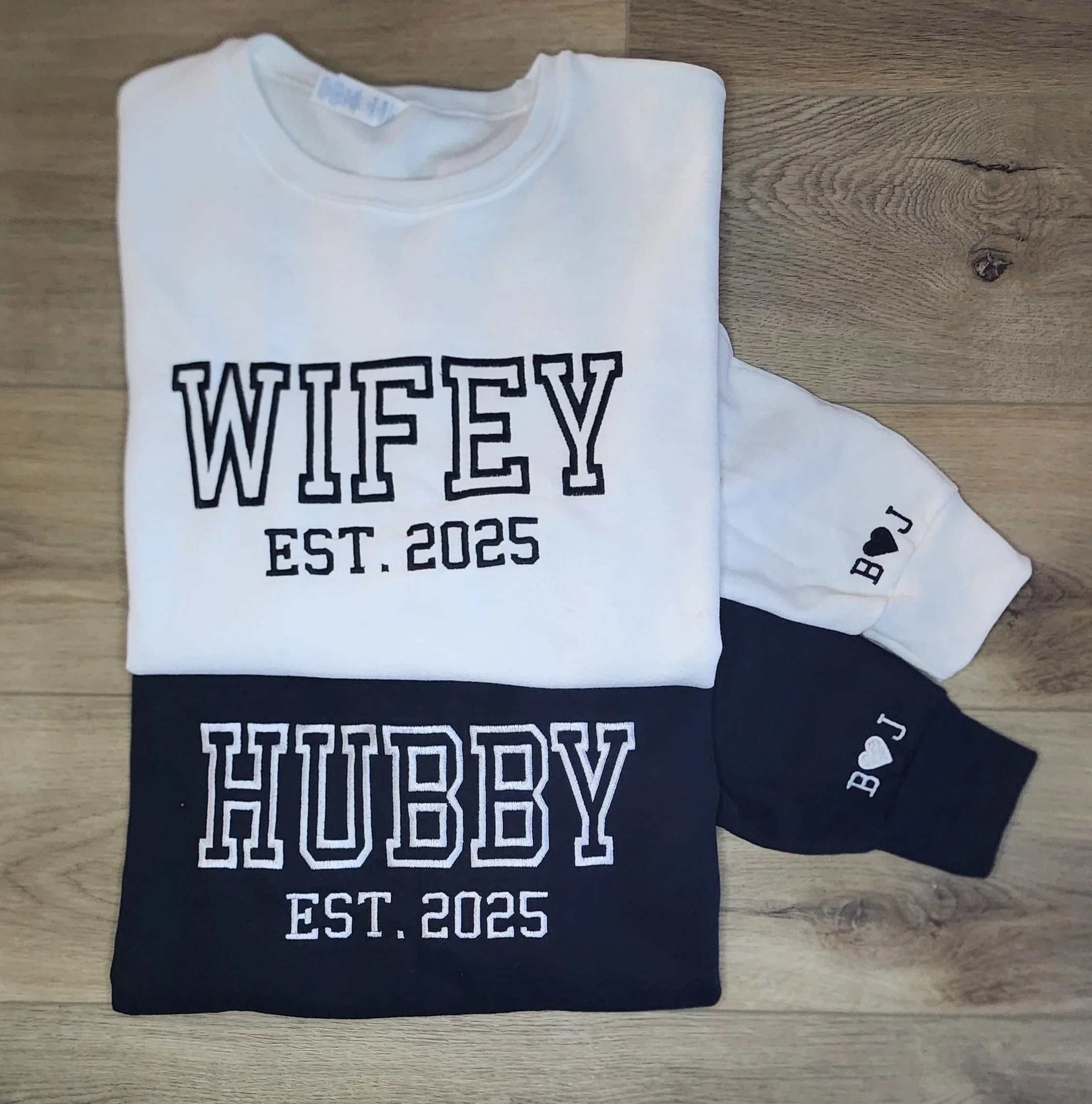 set of white and black sweatshirts with a design embroidered across chest. White one has wifey est. 2025 and black one has hubby est. 2025. Small initials and a heart embroidered on cuffs