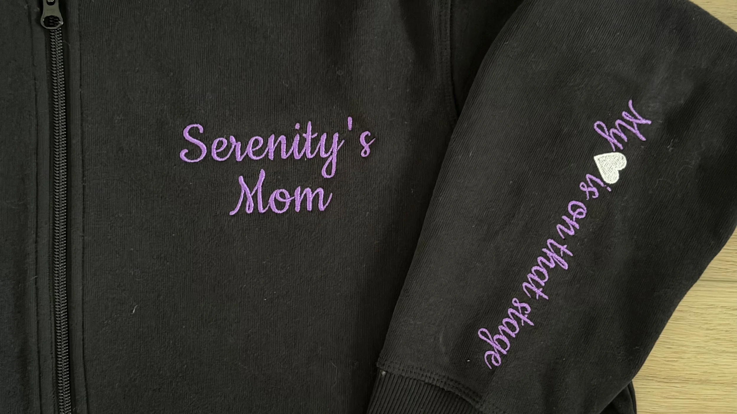 black zip up hoodie with purple text embroidered on a left chest and sleeve