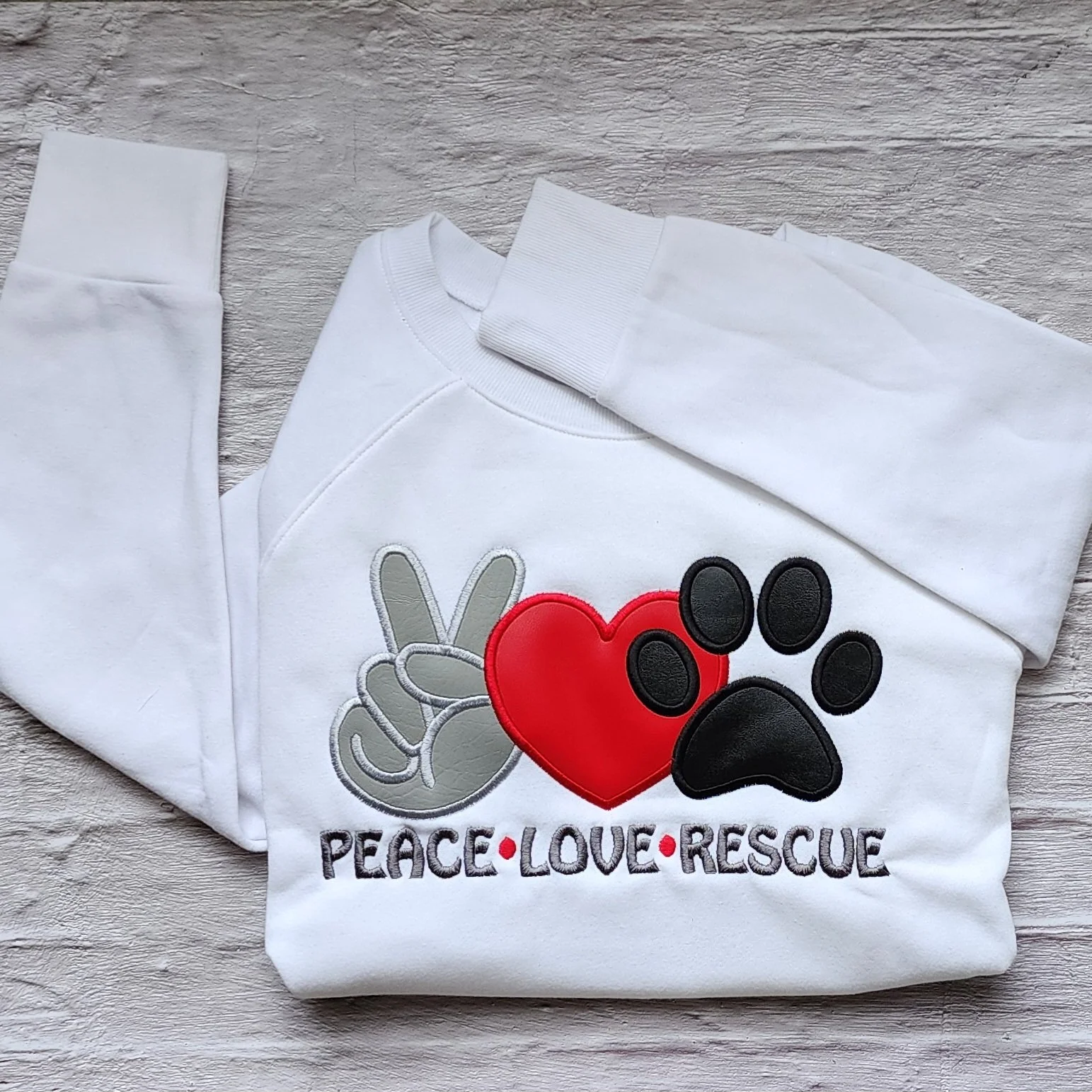 folded white sweatshirt with an applique of a hand making peace sign, heart and dog paw across the chest, underneath embroidered "peace, love, rescue"