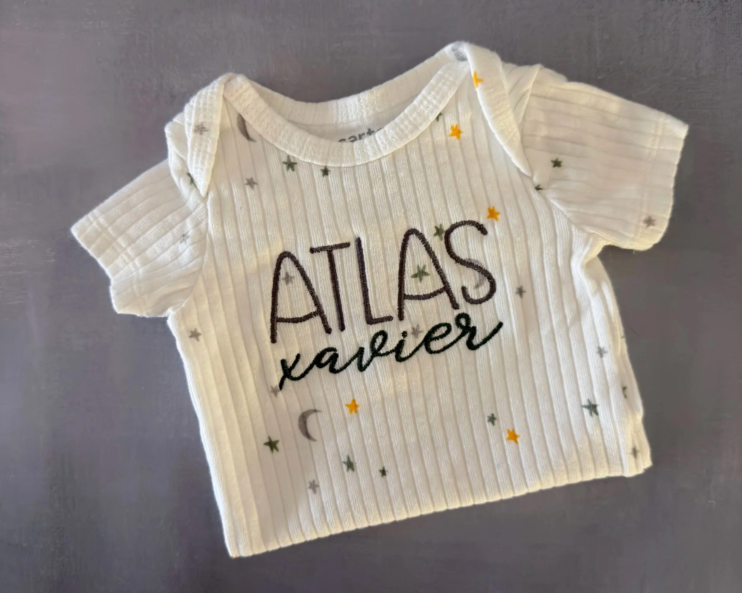 folded white baby bodysuit with small green and yellow stars and moons and embroidered name across the chest, laying on the gray background