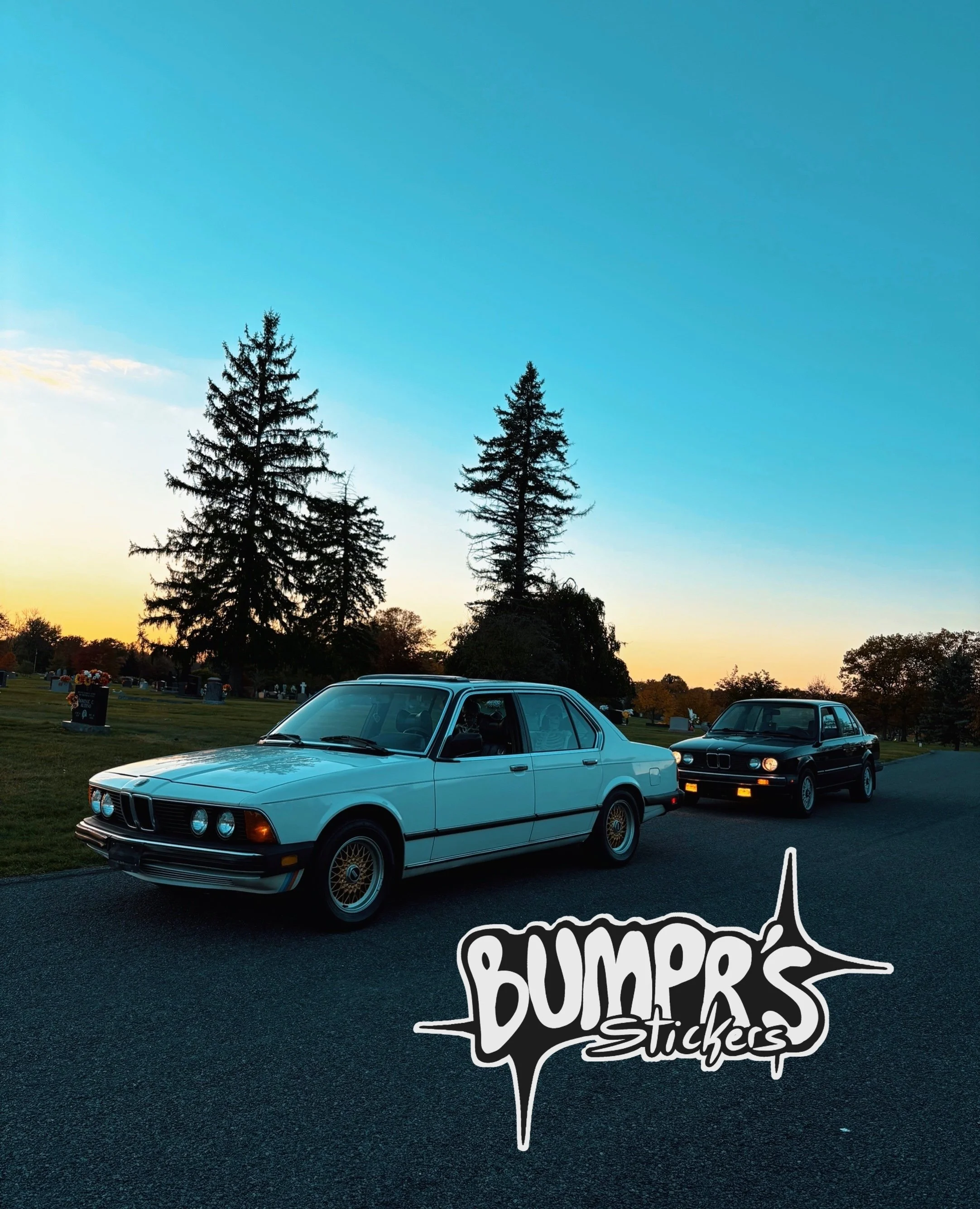 Two classic BMW cars parked on a road in a rural setting during sunset, with trees and gravestones visible in the background, and a Bumper's Stickers logo in the bottom right corner.