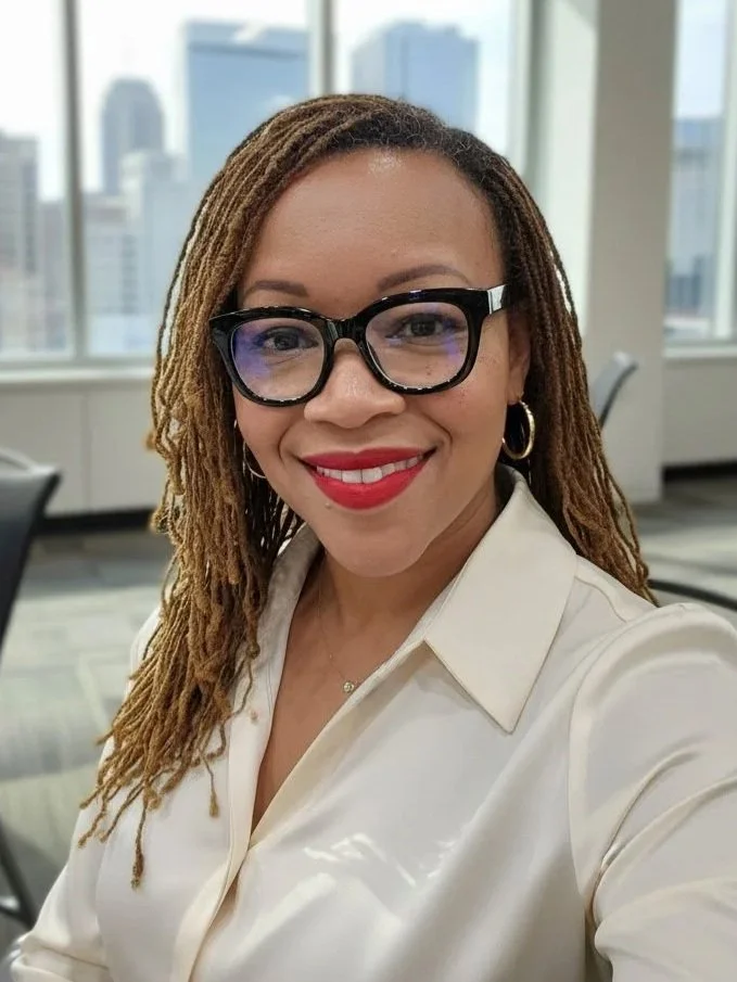 A woman with long dreadlocks, wearing glasses and a white blouse, smiling in a bright office with city buildings visible through large windows.
