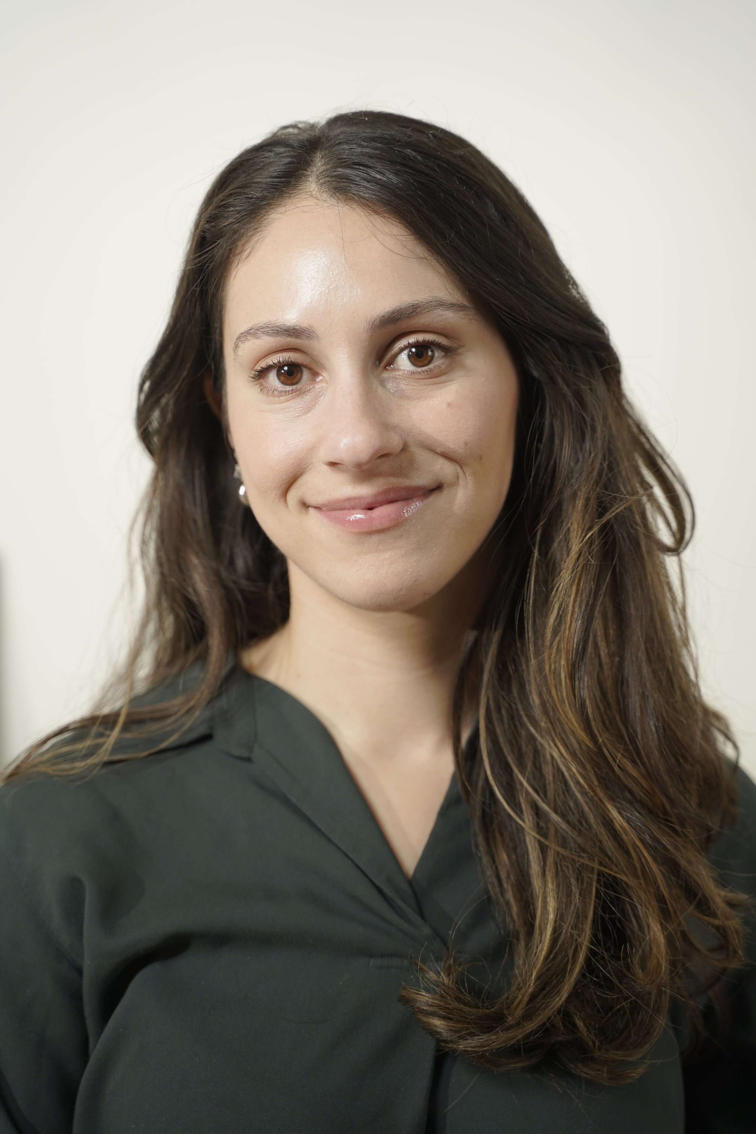 A woman with long dark brown hair and light makeup, wearing a dark green top, smiling softly against a plain light background.