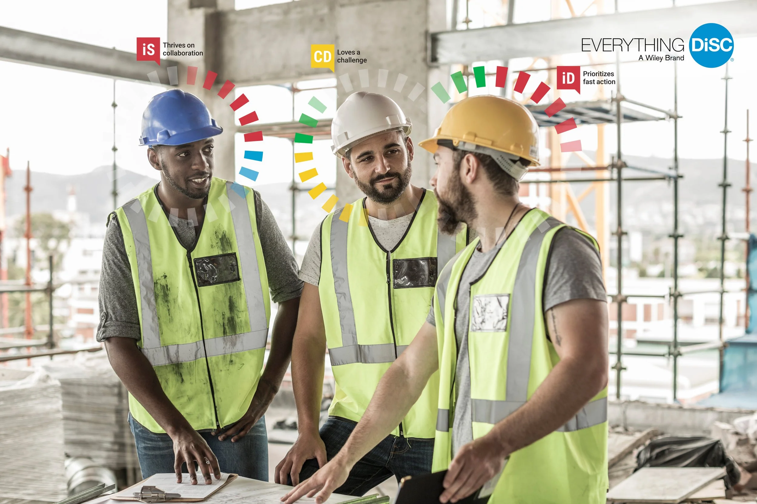 Three construction workers wearing yellow and blue safety vests and hard hats talking on a building site with scaffolding in the background.