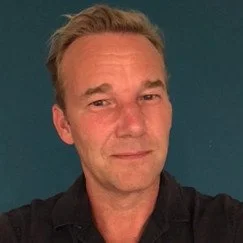 Close-up of a man with short blonde hair, smiling slightly, against a dark teal background.