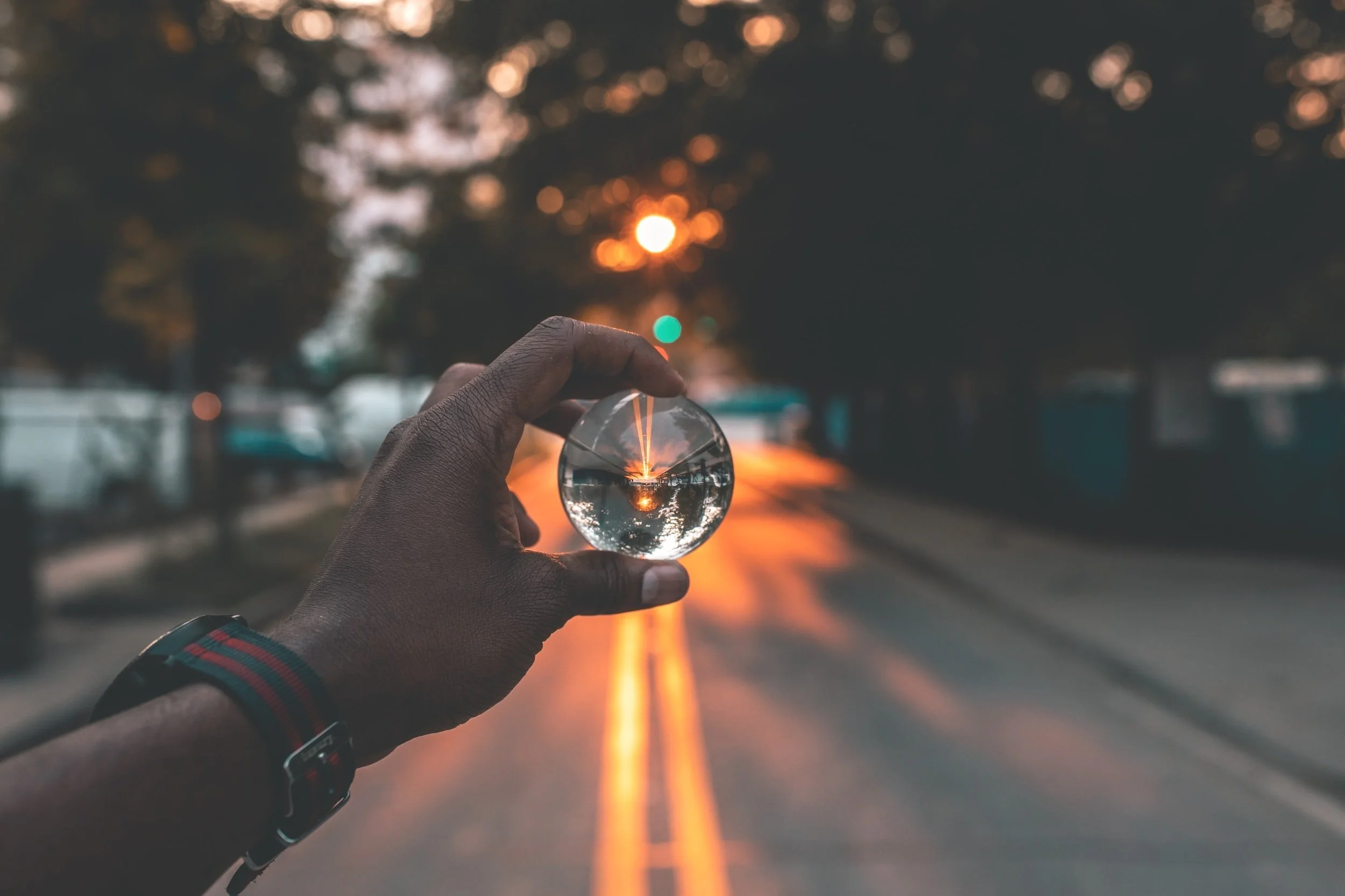 Person holding a glass sphere on a street at sunset, with the sphere reflecting the sunset and street scene upside down.